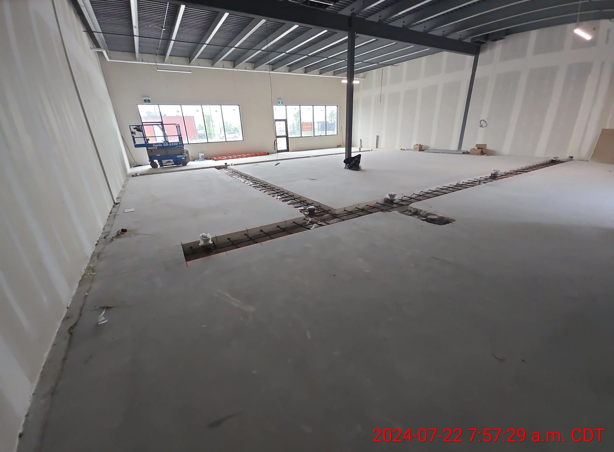 Empty interior space of a building under construction, with visible concrete floor, unfinished walls, and a steel beam in the center, with construction equipment nearby.