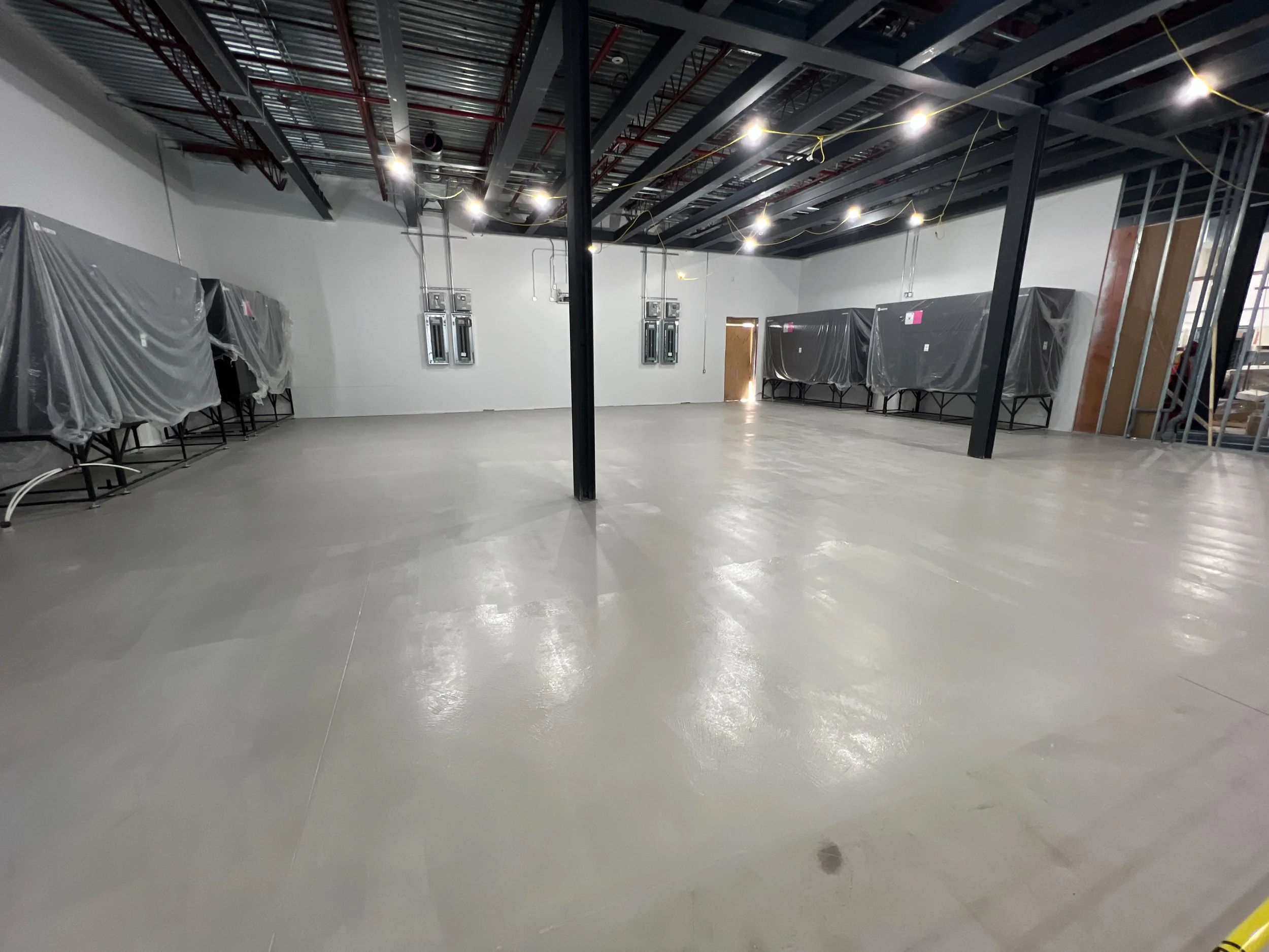Empty construction site with a polished concrete floor and partially finished ceiling with exposed ductwork and wiring. Covered equipment is lined up along the walls, and a staircase is visible on the right side.