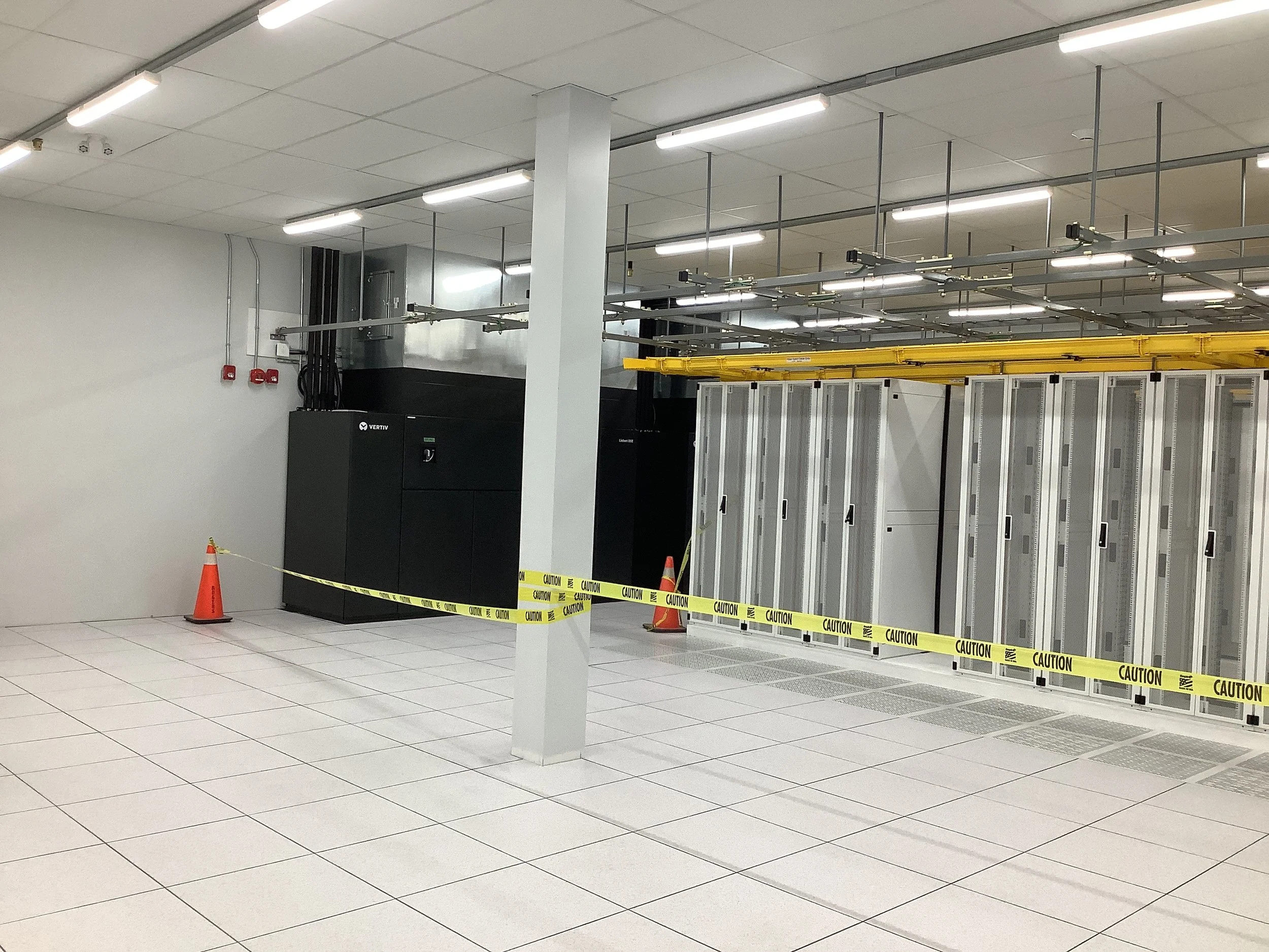 A room with large computer server cabinets, caution tape and cones indicating restricted area, and industrial lighting overhead.