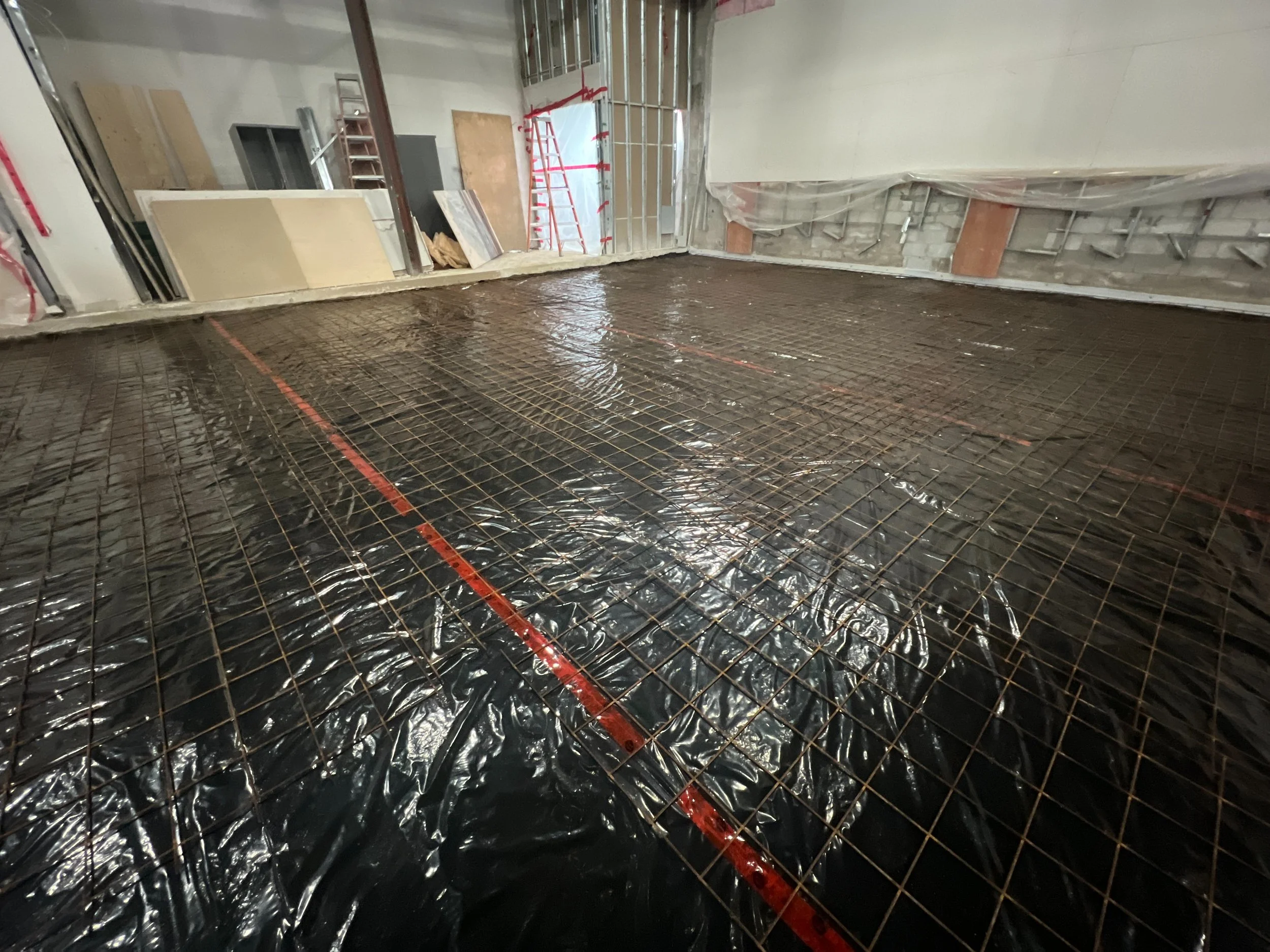 Interior of a building under construction with a concrete floor prepared with rebar and plastic sheeting, construction materials, and ladders in the background.
