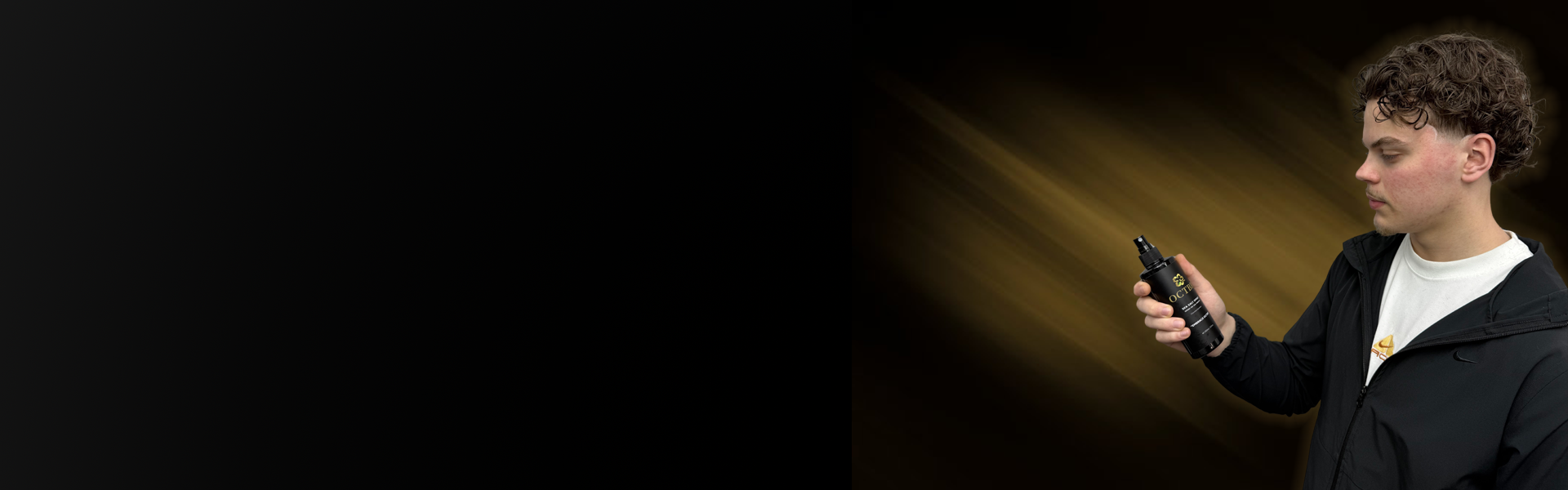 Young man with curly hair wearing a black jacket and white t-shirt, holding a black spray bottle labeled 'OCTBR' with gold accents, against a background with a dark gradient and gold light streaks.