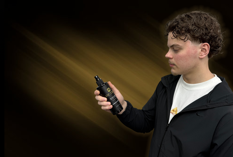 A young man with curly hair looking at a spray bottle labeled 'OCTBR' against a dark background with light streaks.