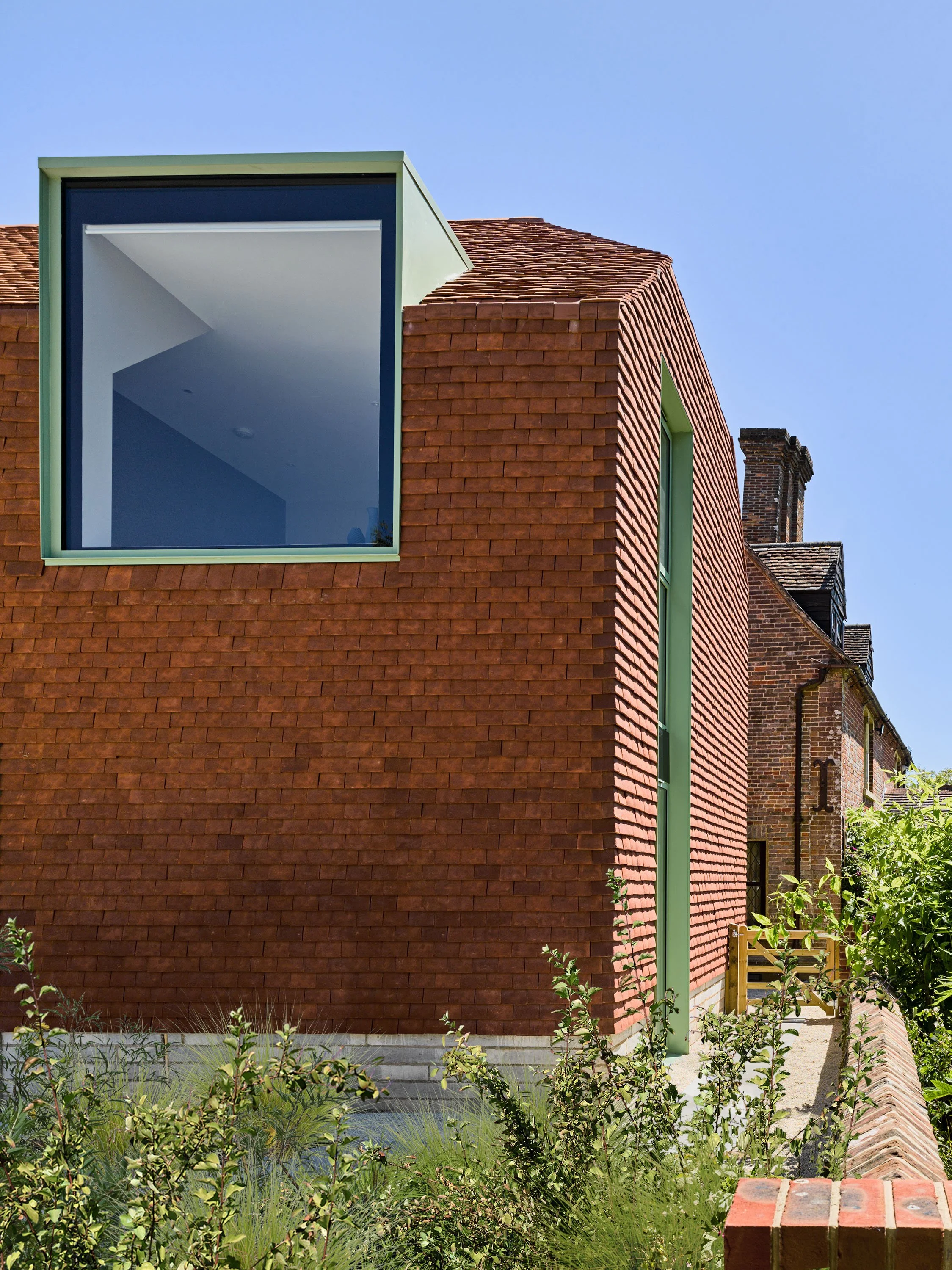 Modern house with red brick exterior, large square window, and green trim, surrounded by plants and garden.