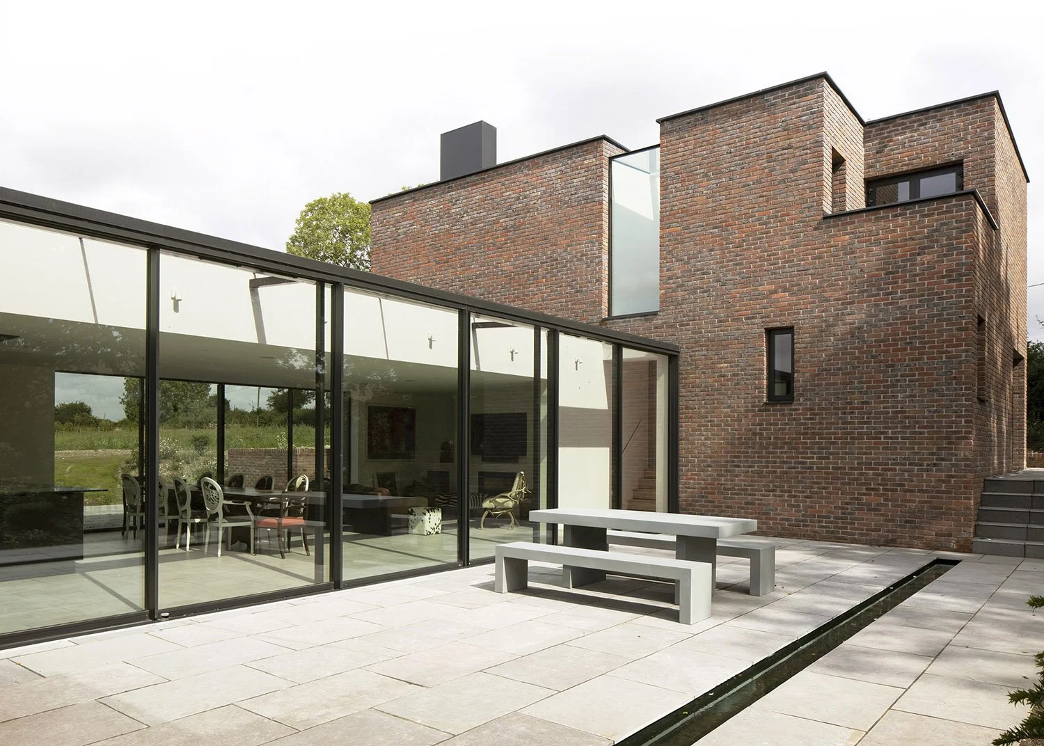 Modern house with a brick exterior and large glass windows, outdoor patio with concrete table and benches, minimalistic design.