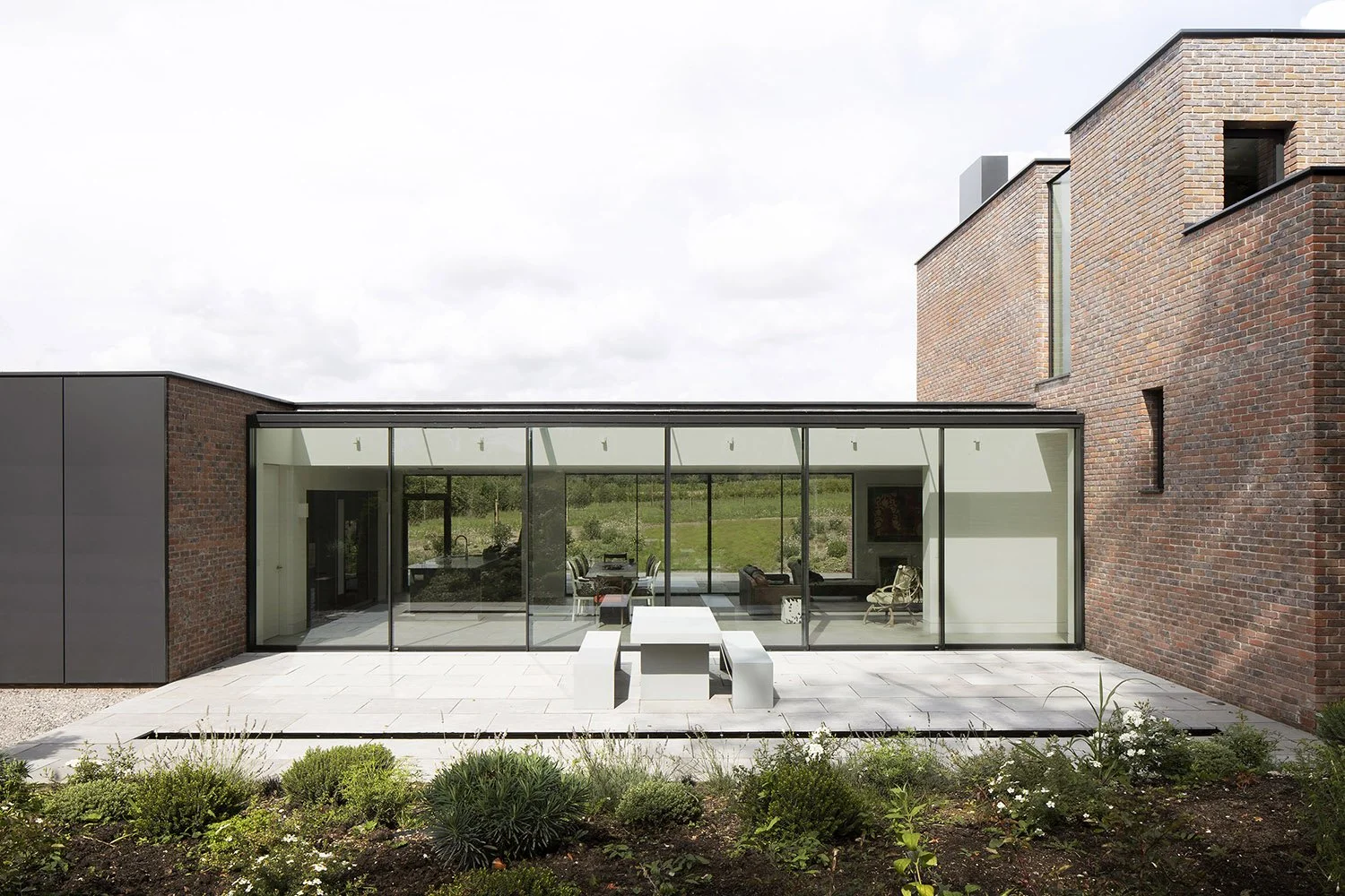 Modern house with large glass windows, brick exterior walls, outdoor patio with white furniture, and landscaped garden in the foreground.