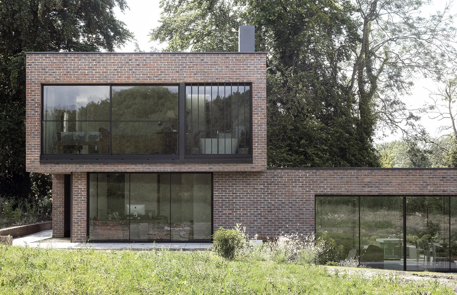 Modern brick house with large glass windows in a grassy yard, surrounded by trees.