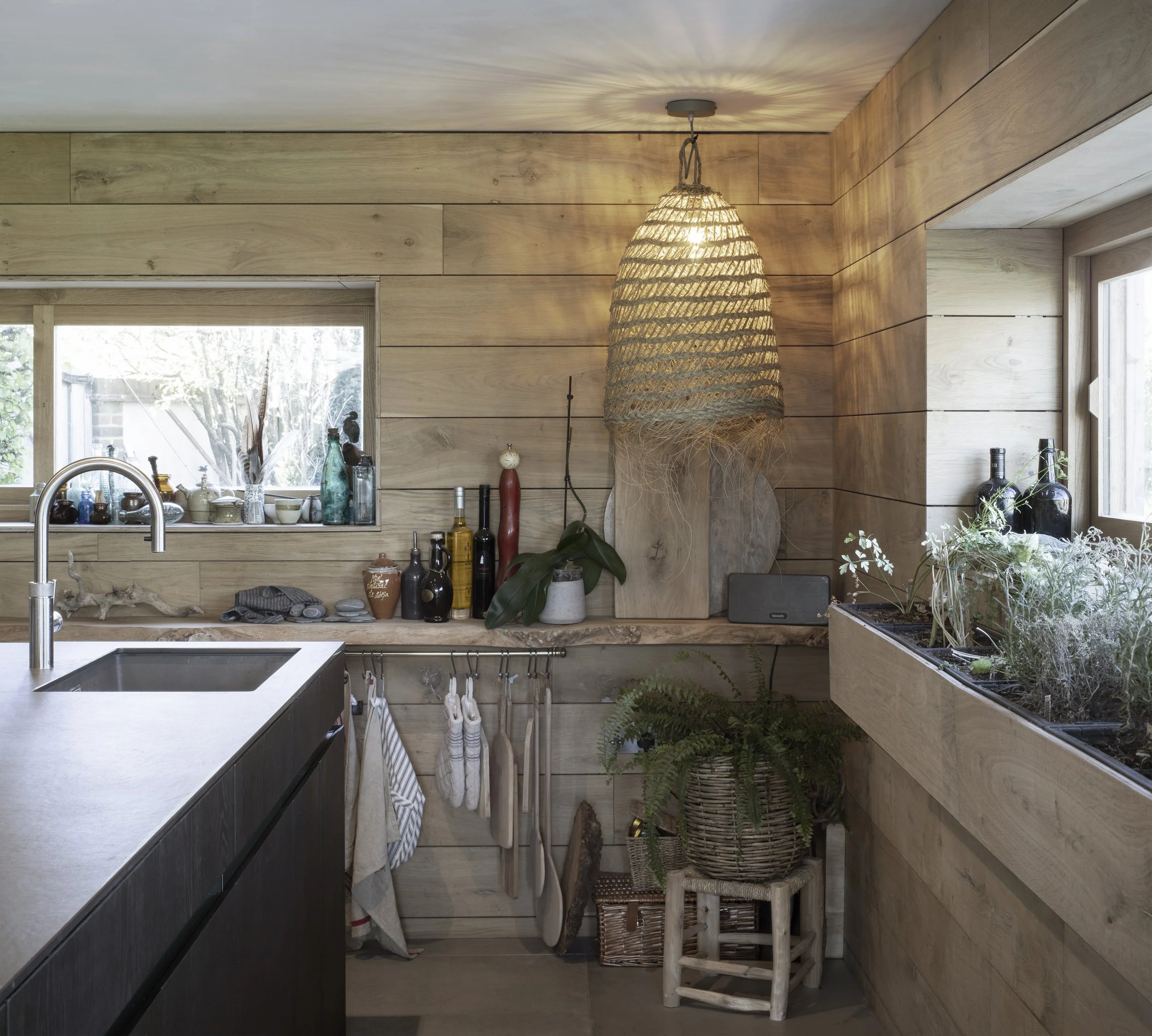 A rustic kitchen with wooden panel walls, a window above the sink, and a hanging woven light fixture. The counter has bottles, plants, and kitchen utensils, with a small stool holding a potted fern and a basket underneath. There is a window with a vi