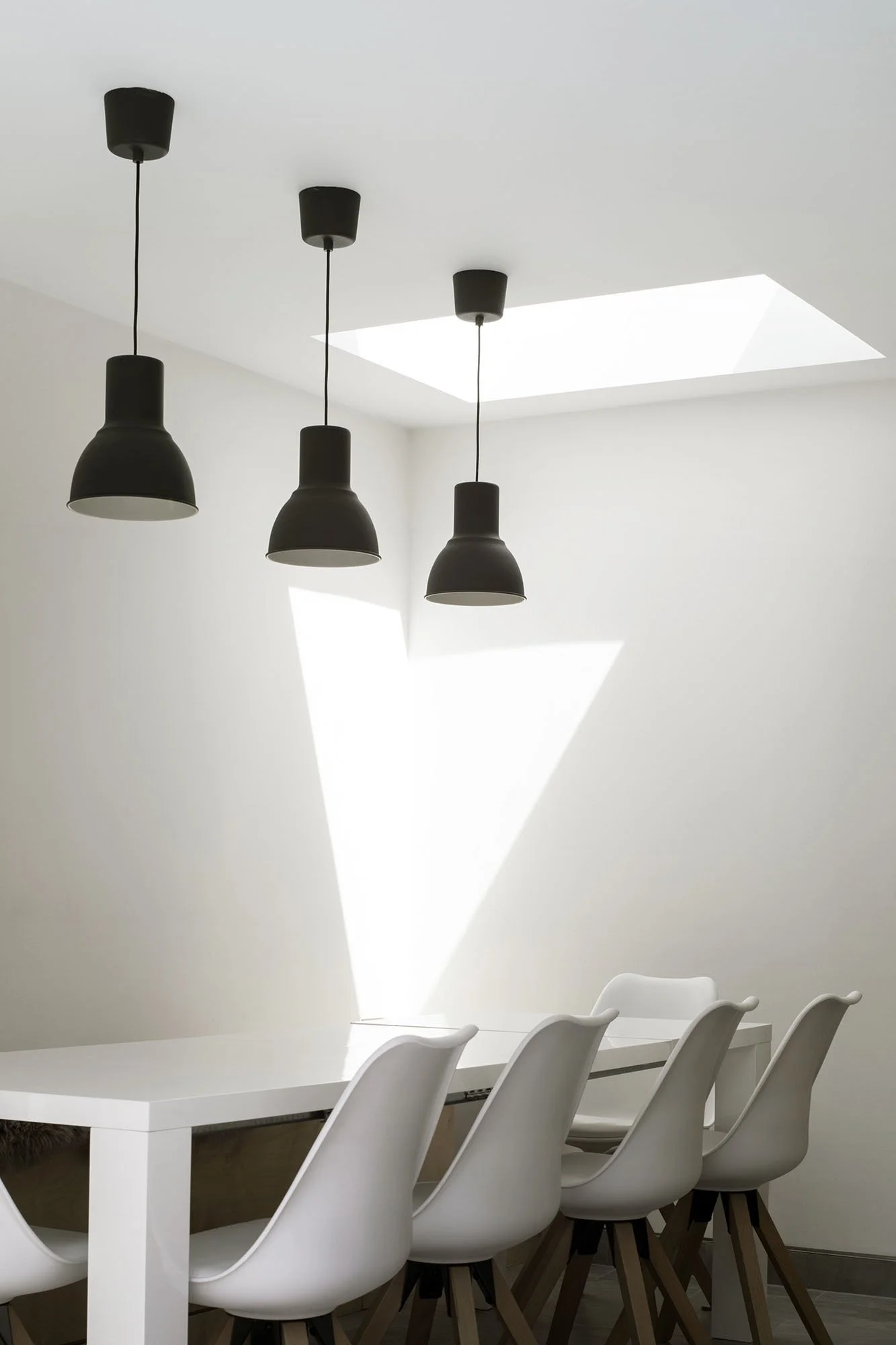 Modern dining room with white chairs around a white table, black pendant lights hanging from ceiling, and a skylight letting in natural light.