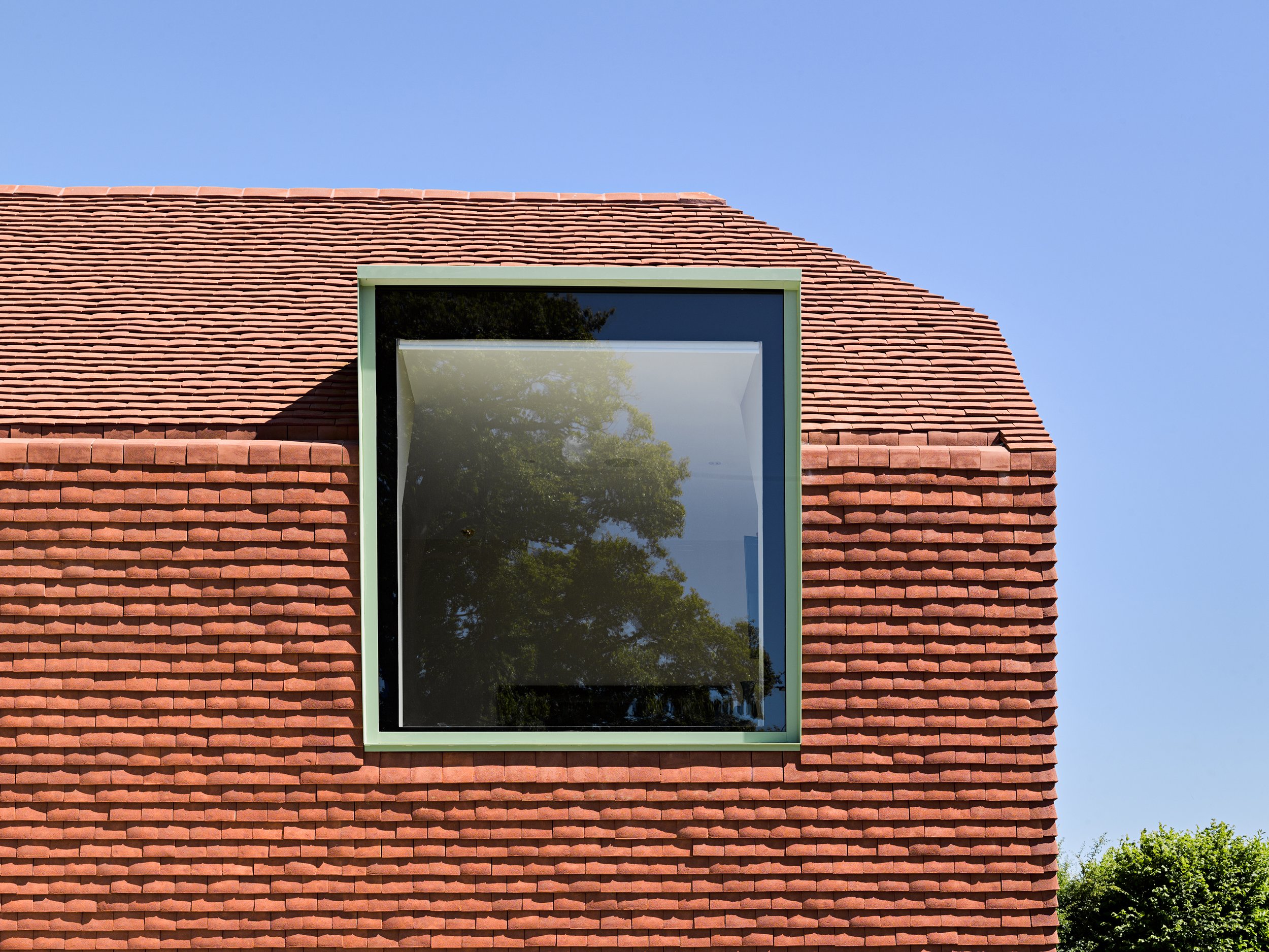 Close-up of a modern building's exterior with red brick siding and a large square window reflecting trees and a blue sky.