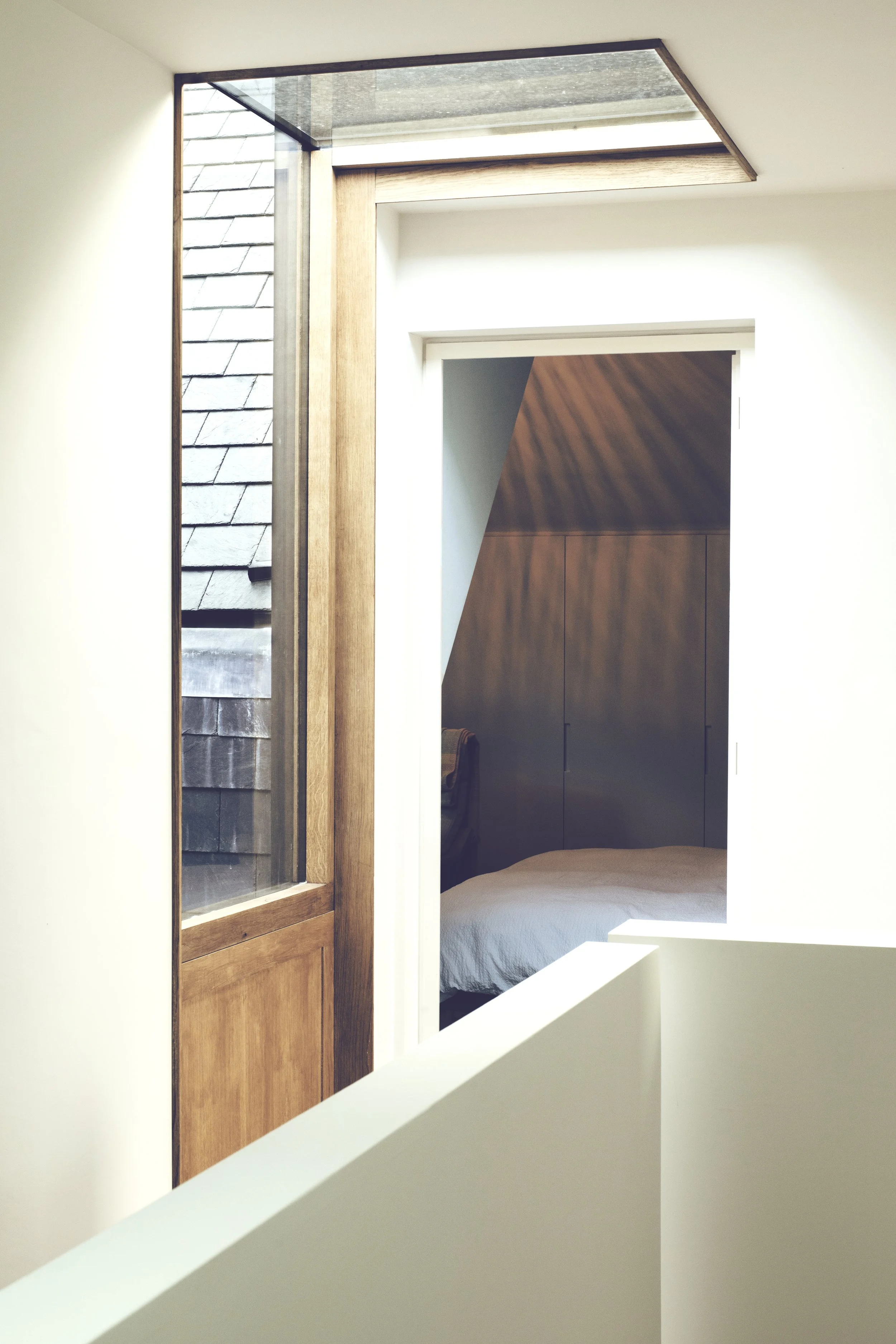 A view through an open doorway from a white corridor into a bedroom with a built-in wooden wardrobe and a bed with a beige duvet.