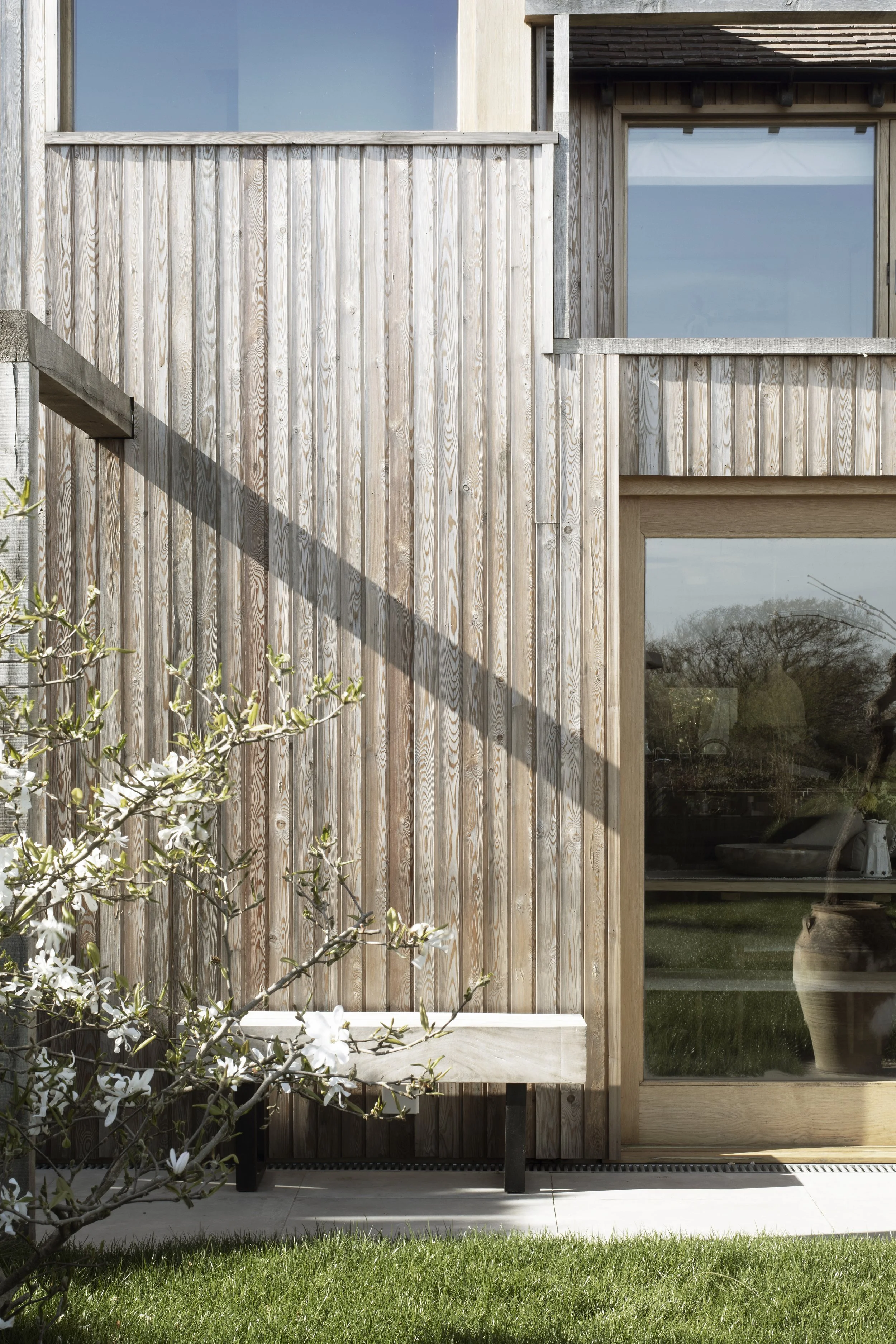 Modern house with wooden exterior and large glass windows, outdoor bench, flowering shrub, and grassy lawn.