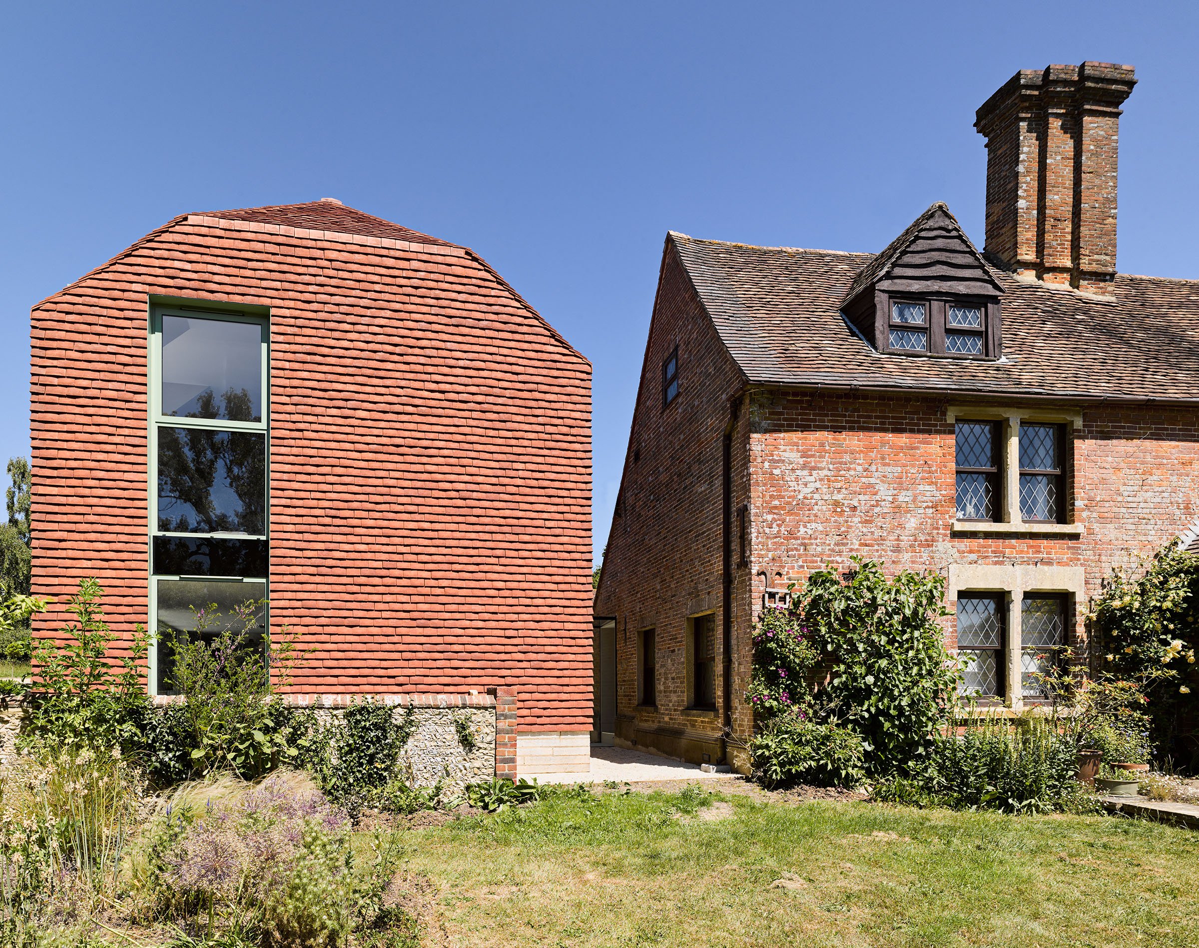Two contrasting houses side by side: one with modern architecture, red brick exterior, and large window, and the other with traditional brick design, small diamond-pane windows, and a chimney, set in a garden with greenery under a clear blue sky.