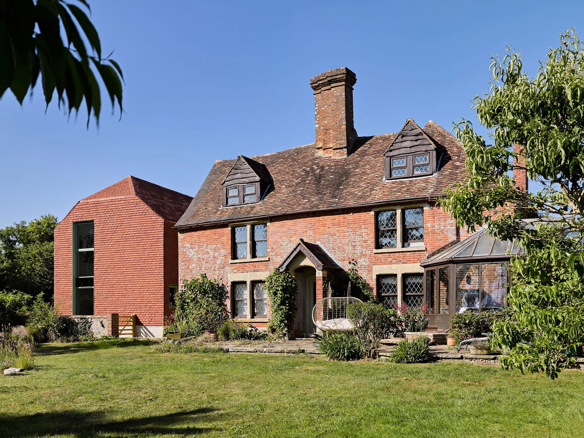 A historic brick cottage with a chimney and dormer windows, alongside a modern red brick addition, surrounded by a green garden with trees and bushes, under a clear blue sky.