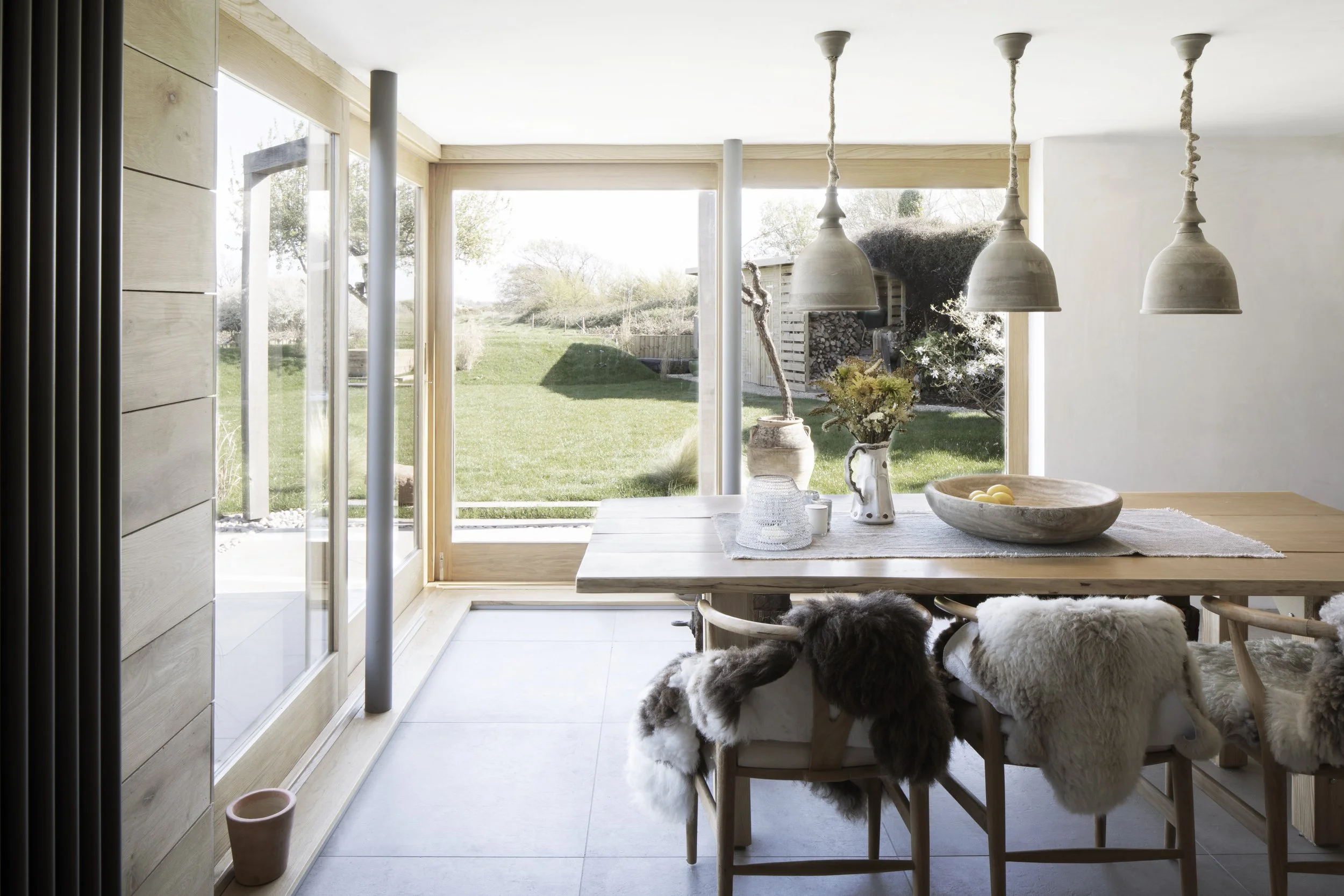 Bright dining room with large glass doors opening to a grassy backyard, wooden table decorated with flowers and bowls, and chairs with fur throws.
