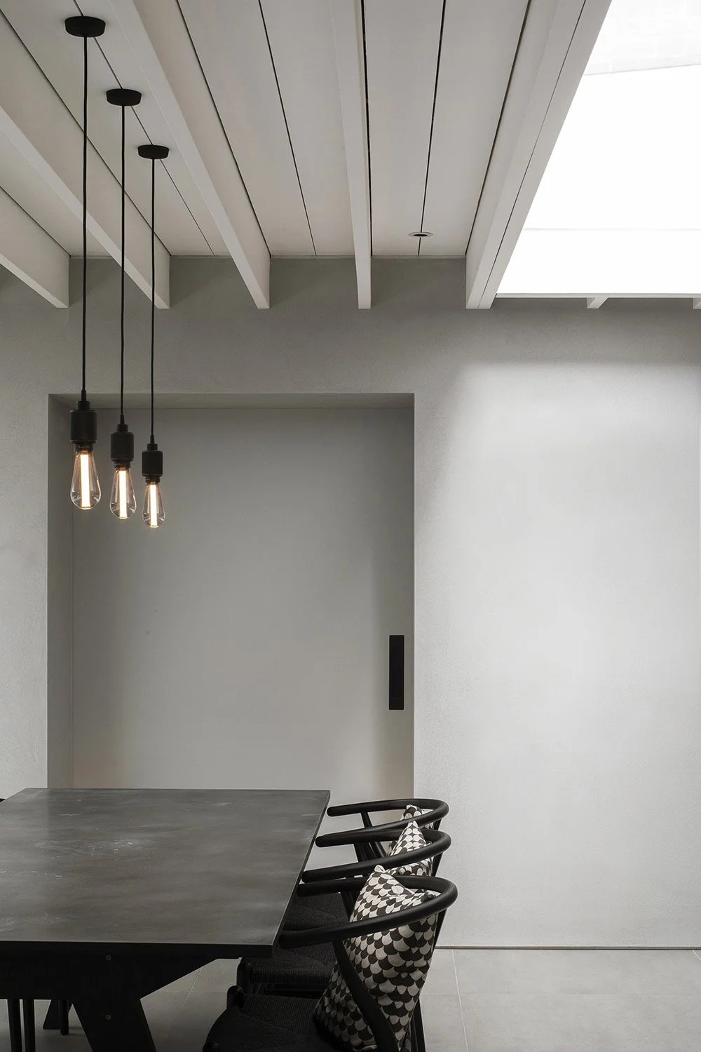 Minimalist dining area with a concrete table, three black chairs with patterned cushions, three Edison-style pendant lights hanging from the ceiling, and a high ceiling with white-paneled beams and a skylight.