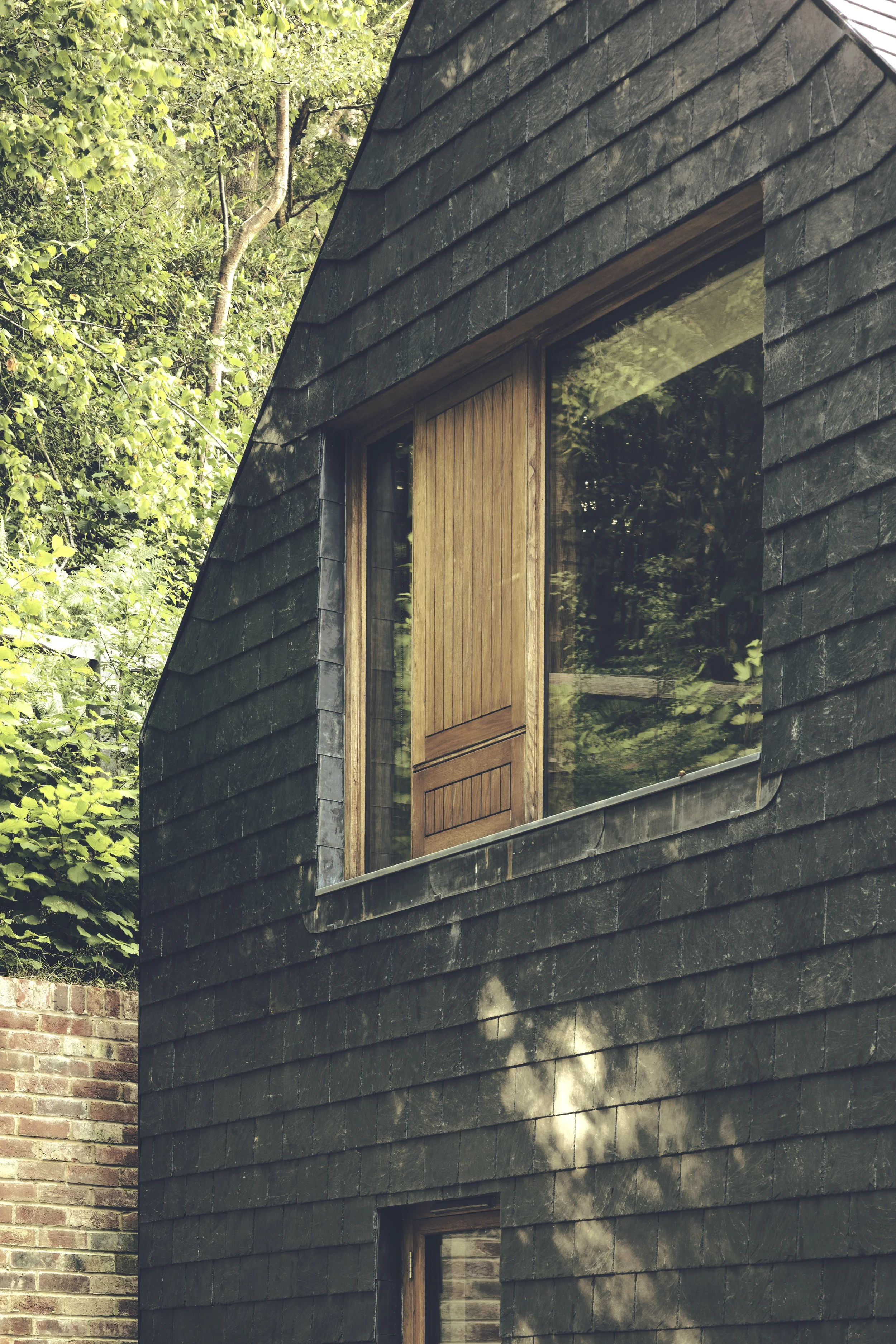 Close-up of a modern black brick house with a large window and a wooden door, surrounded by green trees and a brick wall in the background.