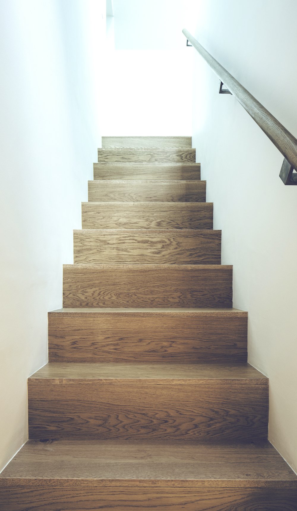 Wooden staircase with a handrail on the right side, leading upward towards a bright light.
