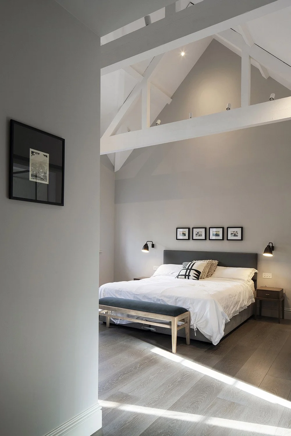 A modern, minimalist bedroom with a white bed, black wall-mounted lamps, a framed picture above the headboard, and artwork on the side wall, with an open ceiling showing exposed beams.