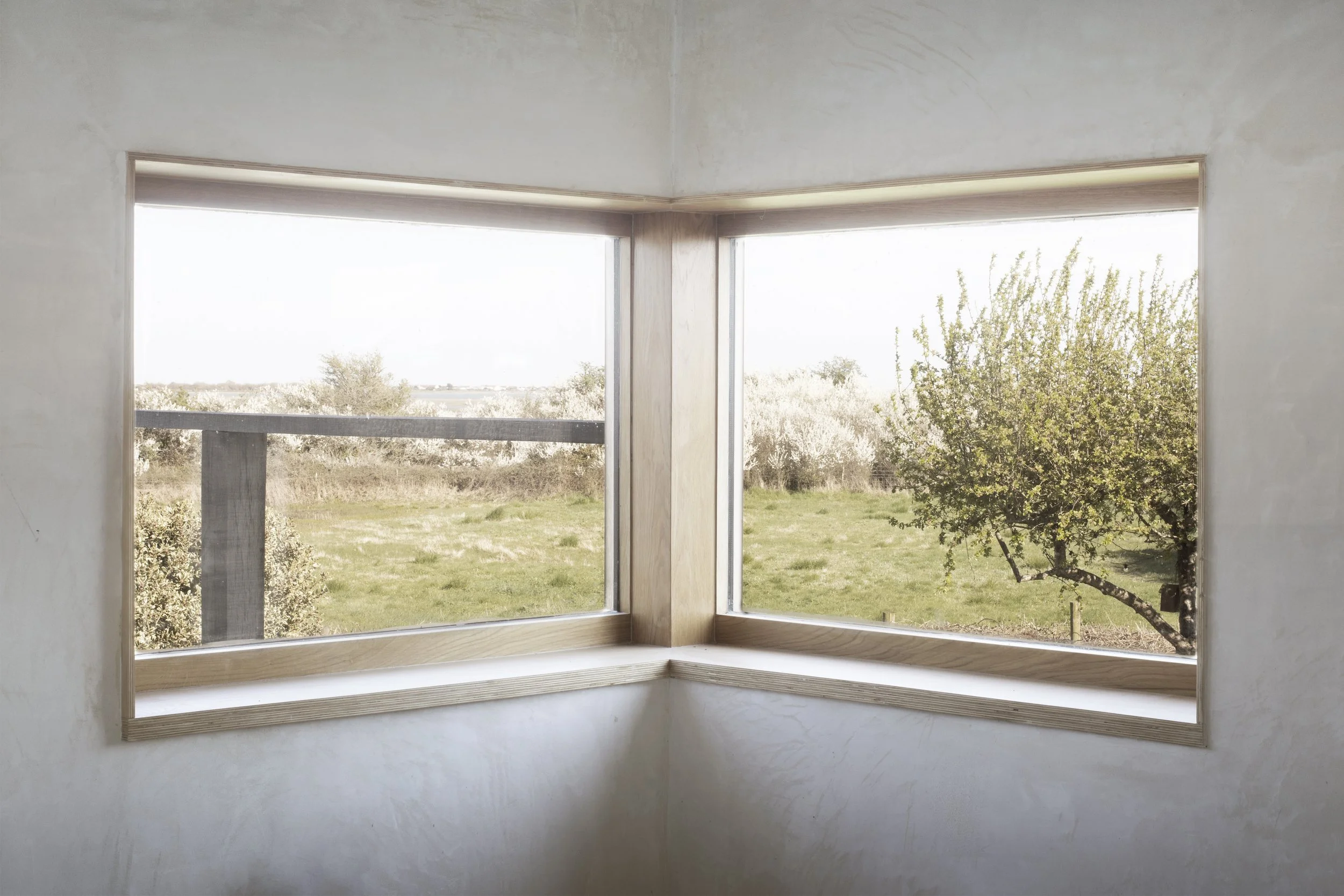 A corner of a room with two large windows showing an outdoor landscape of a grassy field, trees, and a partly cloudy sky.