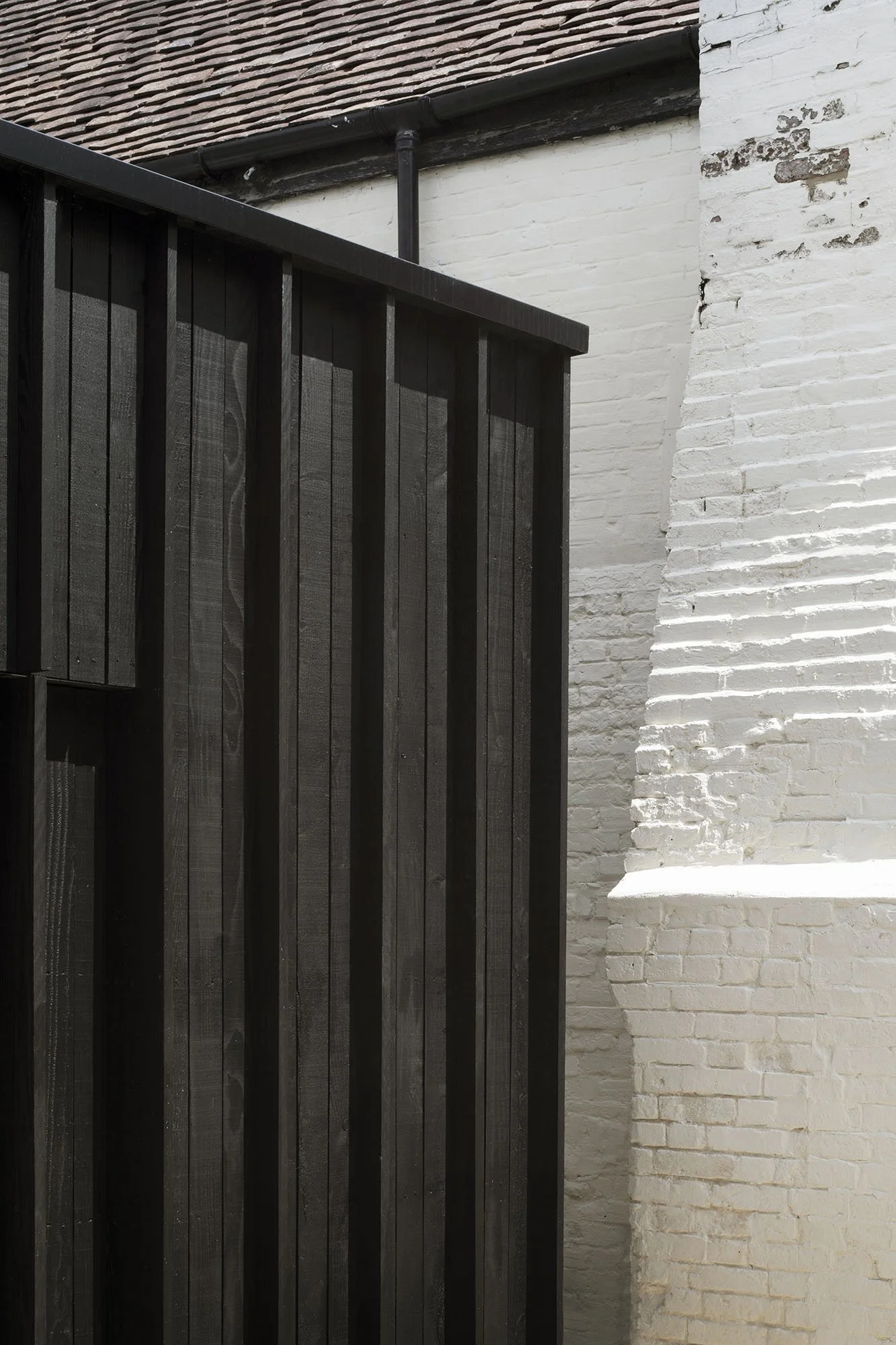 Close-up of a black vertical slat fence against a white brick wall, with a black pipe running above.