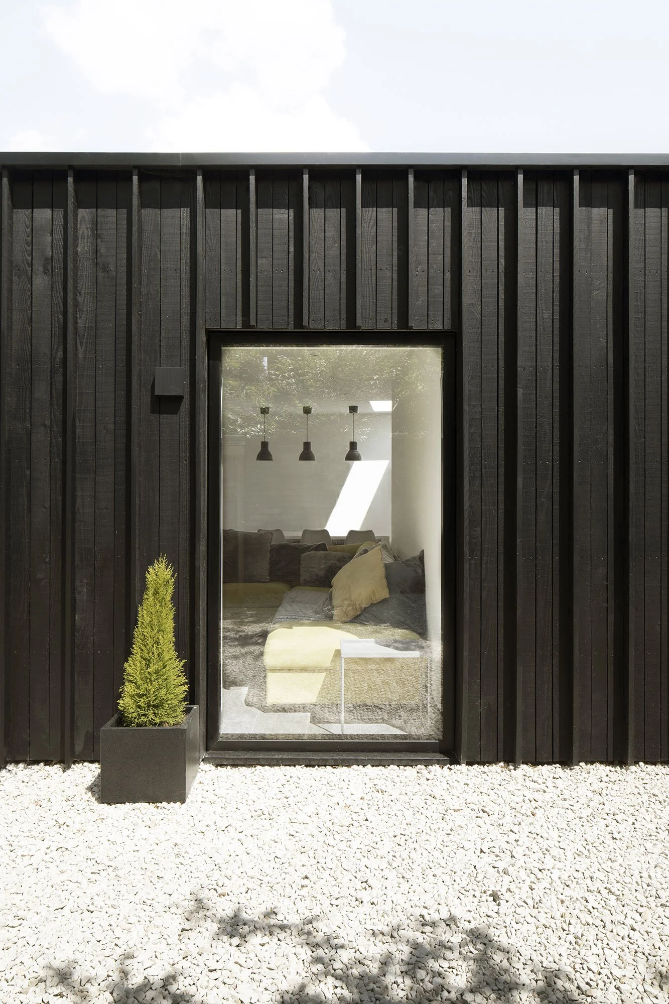 View of a modern black wooden house exterior with a glass door and a potted plant on white gravel outside, reflecting a room with couches, pillows, and hanging pendant lights inside.