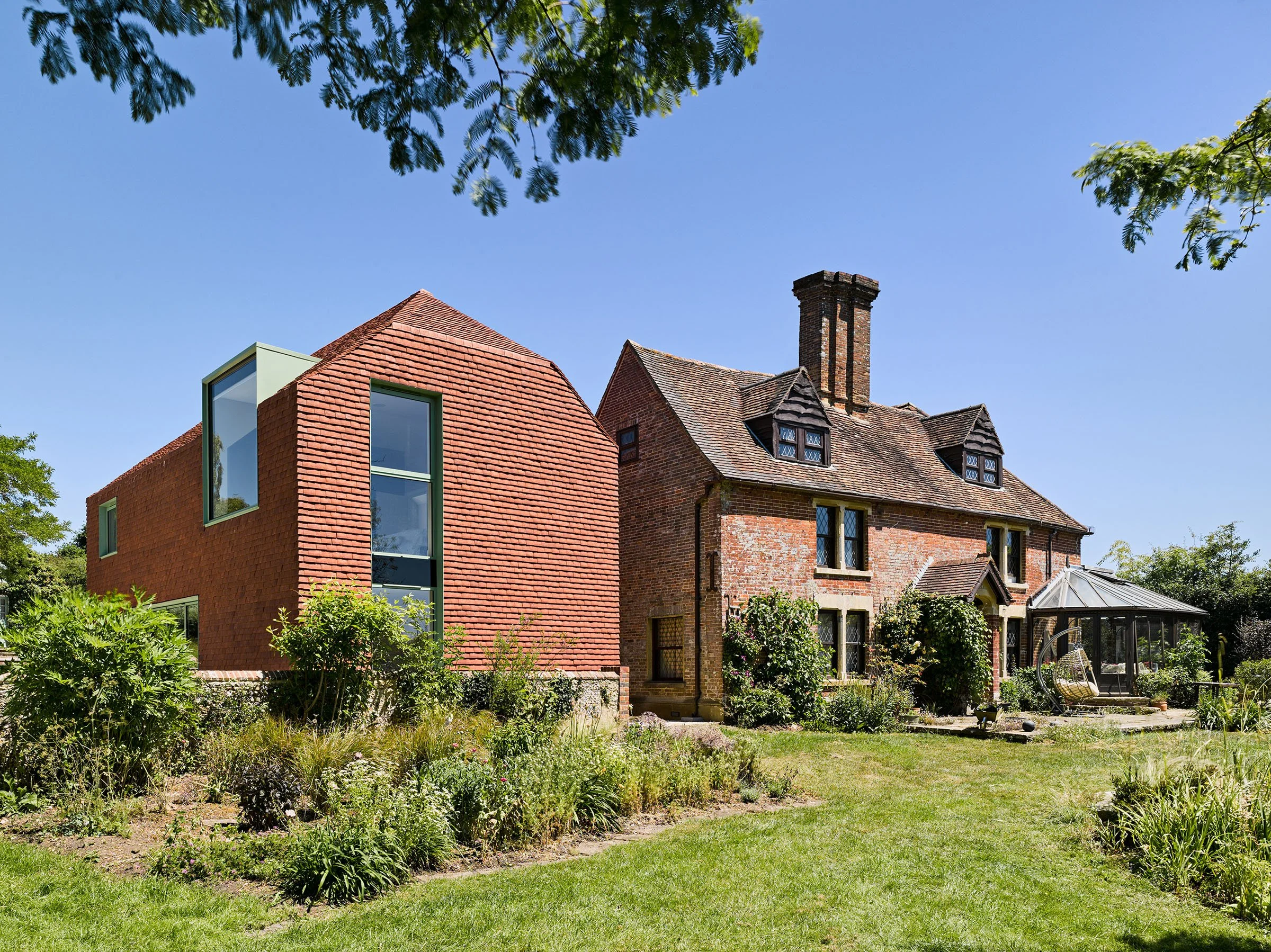 A traditional brick house with a steep gable roof and a modern addition with glass windows, situated in a lush garden with green grass, shrubs, and trees under a clear blue sky.