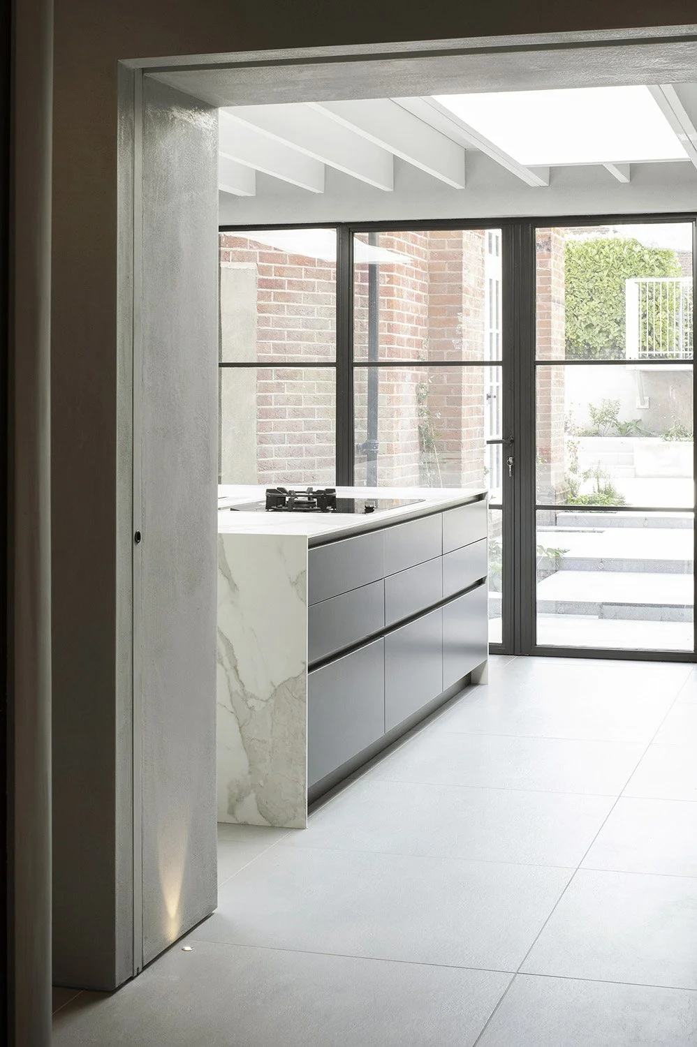 Modern kitchen with minimalist design featuring a marble countertop, gray drawers, and large glass sliding doors leading to an outdoor area with brick walls and greenery.