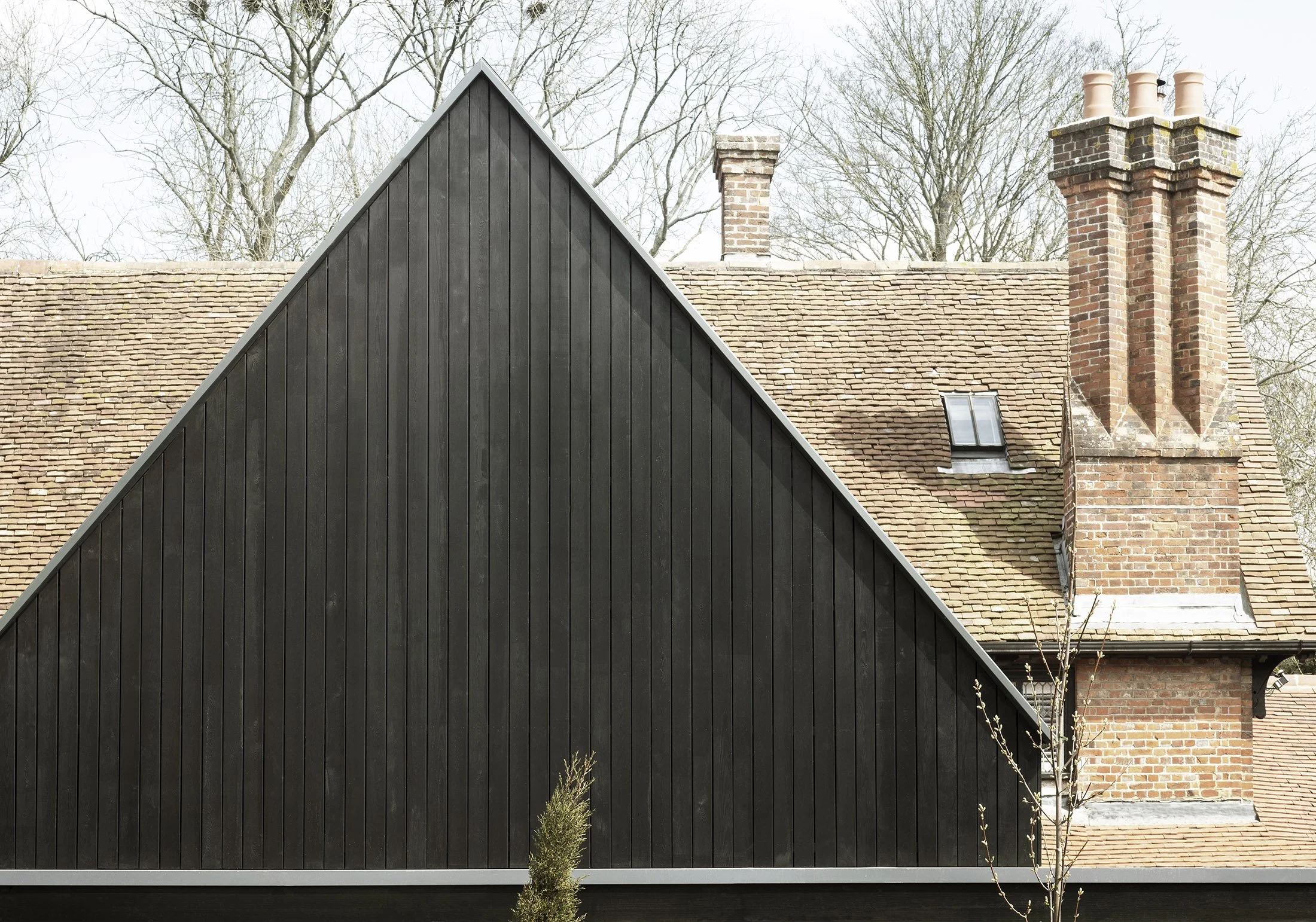 A house with a traditional brick chimney and roof, partially covered by a black modern triangular extension with vertical wood siding, and some trees in the background.