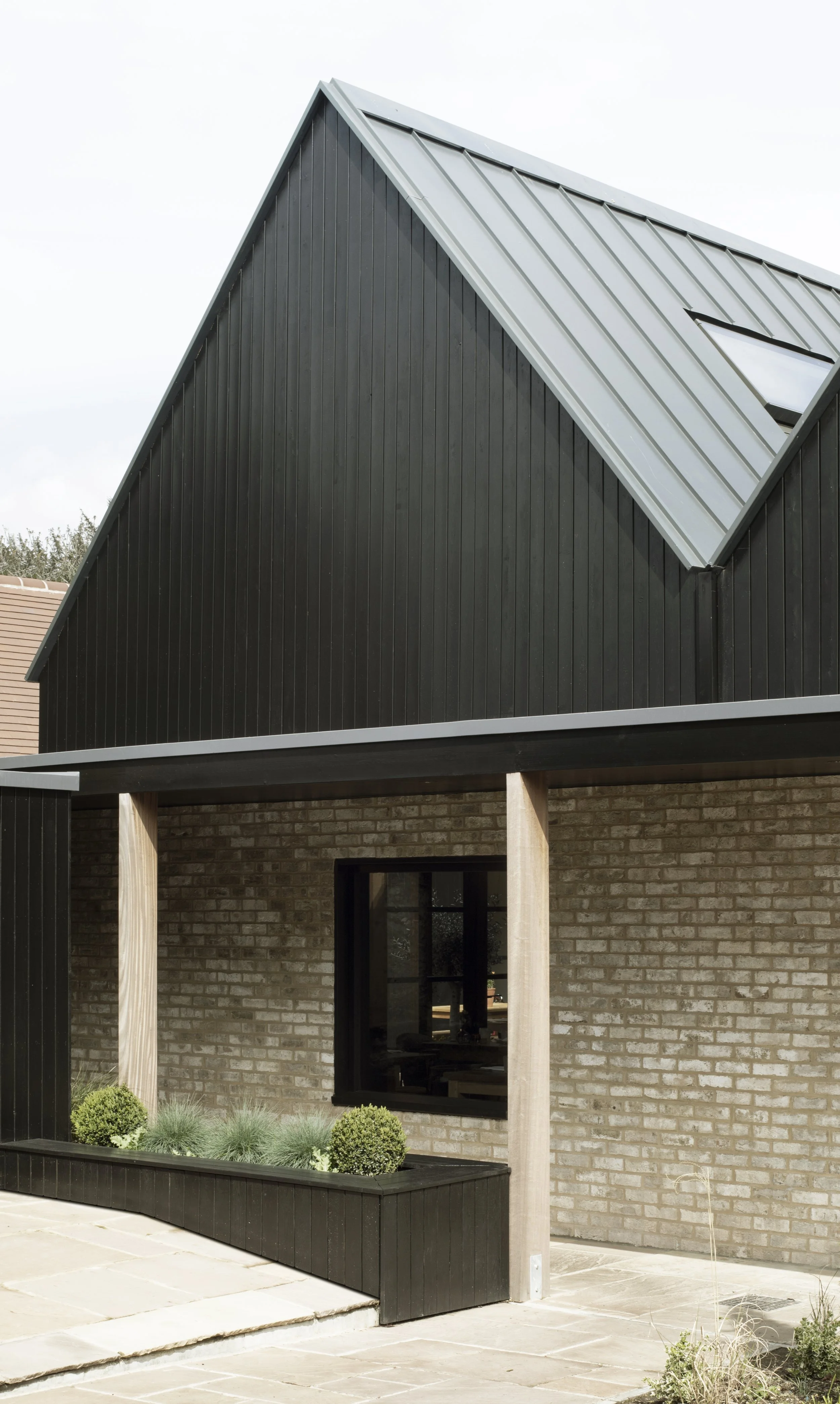 Modern house with black vertical siding, brick lower level, large window, and planters with green shrubs and grasses.