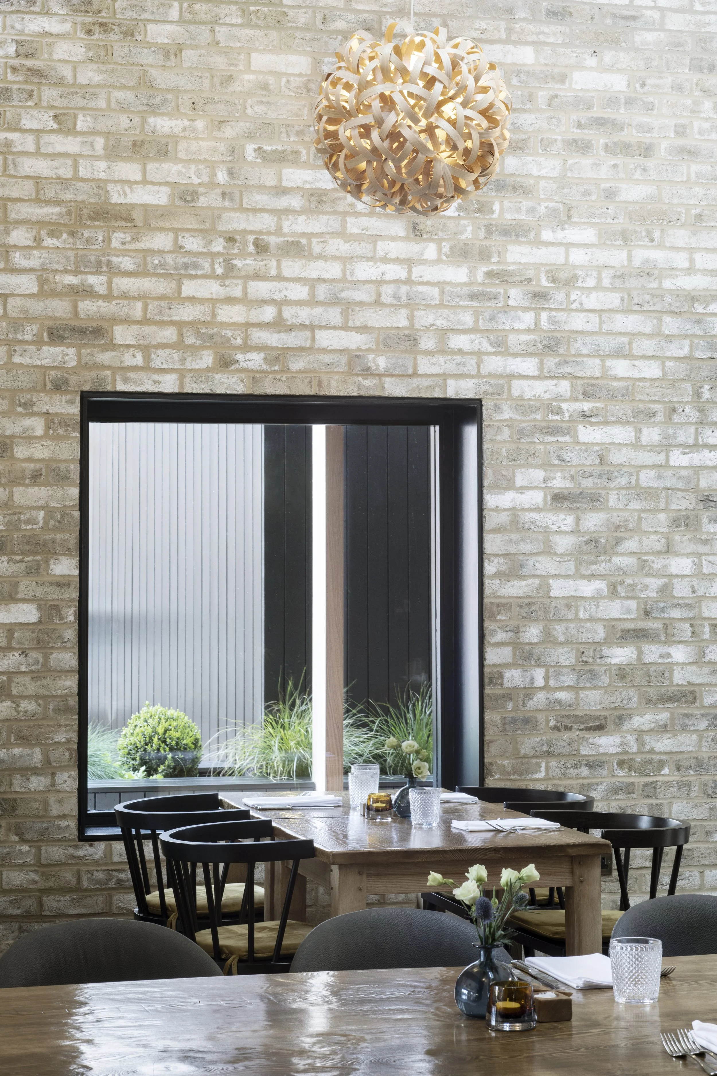 Interior view of a dining area with wooden tables, black chairs, a window showing plants outside, and a modern pendant light hanging from a white brick wall.