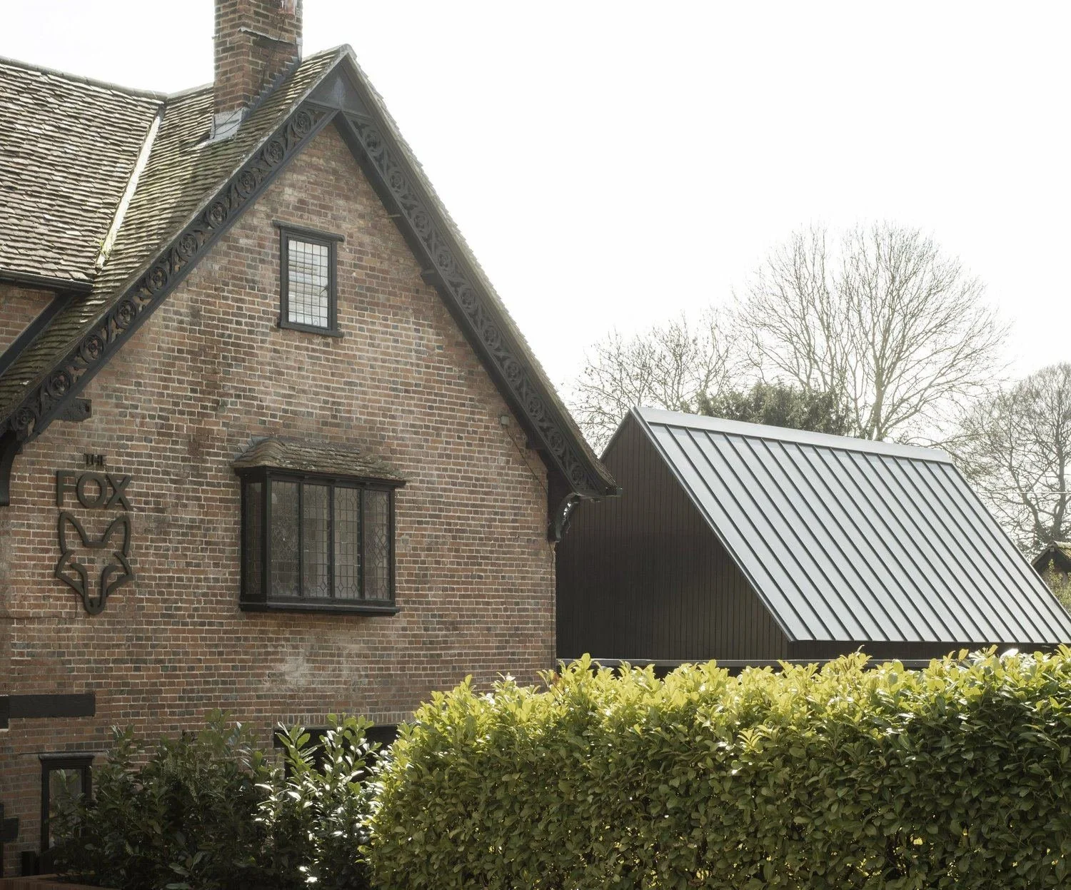 Paul Cashin Architects : A brick house with black trim and a decorative fox sign on the wall, next to a modern metal roof addition, and surrounded by a hedge and leafless trees in the background.