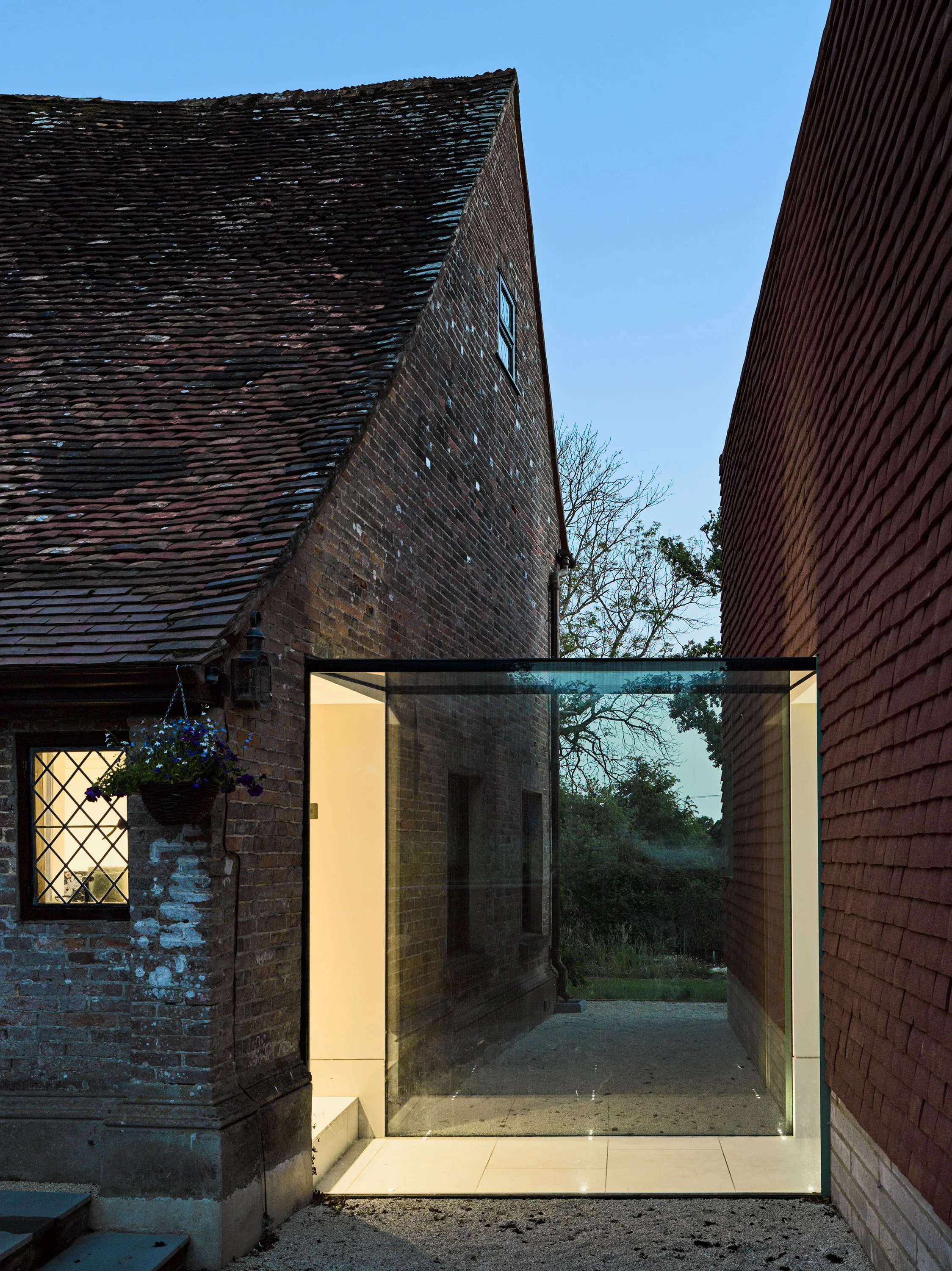 A modern glass entranceway between two traditional brick buildings, with a small outdoor area and trees in the background, during evening with the sky turning blue.