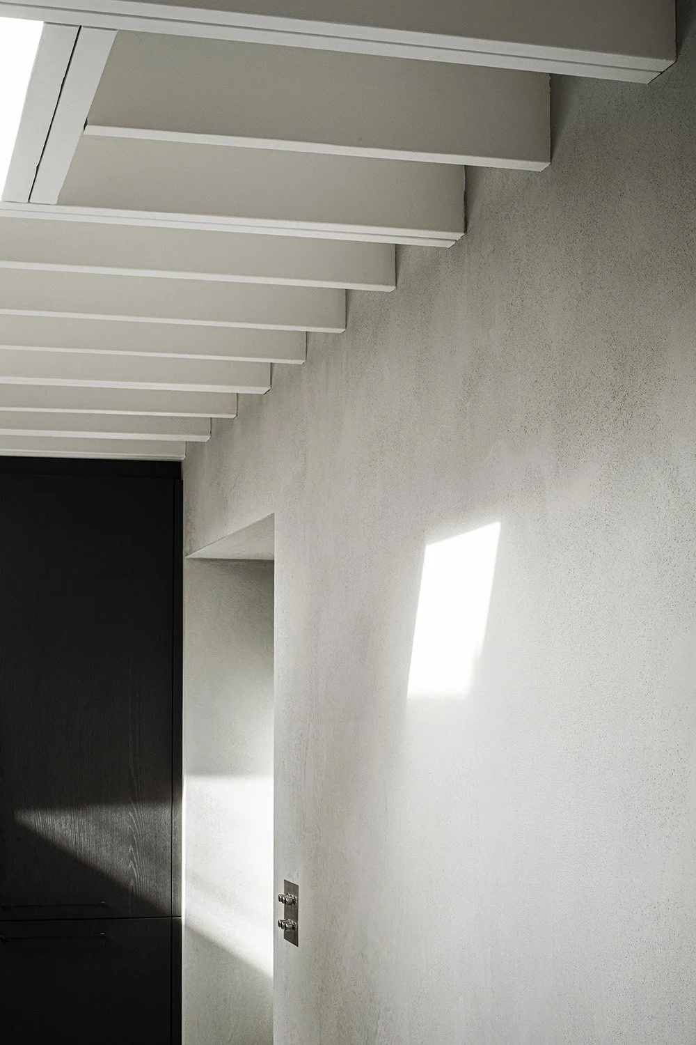 Interior wall with white textured surface, shadow of a window on the wall, black door and horizontal ceiling beams