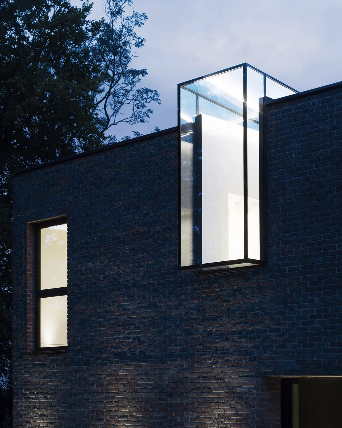 Modern brick building with large glass window on upper floor, illuminated interior, and a tall tree in the background at dusk.