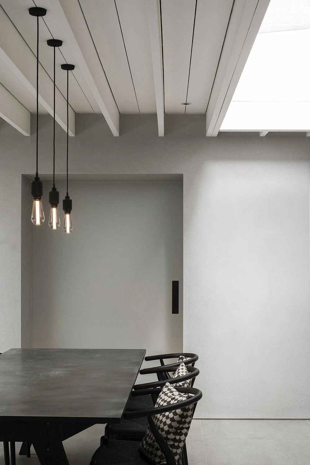 Paul Cashin Architects : Minimalist dining area with gray walls, a dark gray table, black chairs with patterned cushions, and three hanging Edison-style light bulbs from the ceiling.