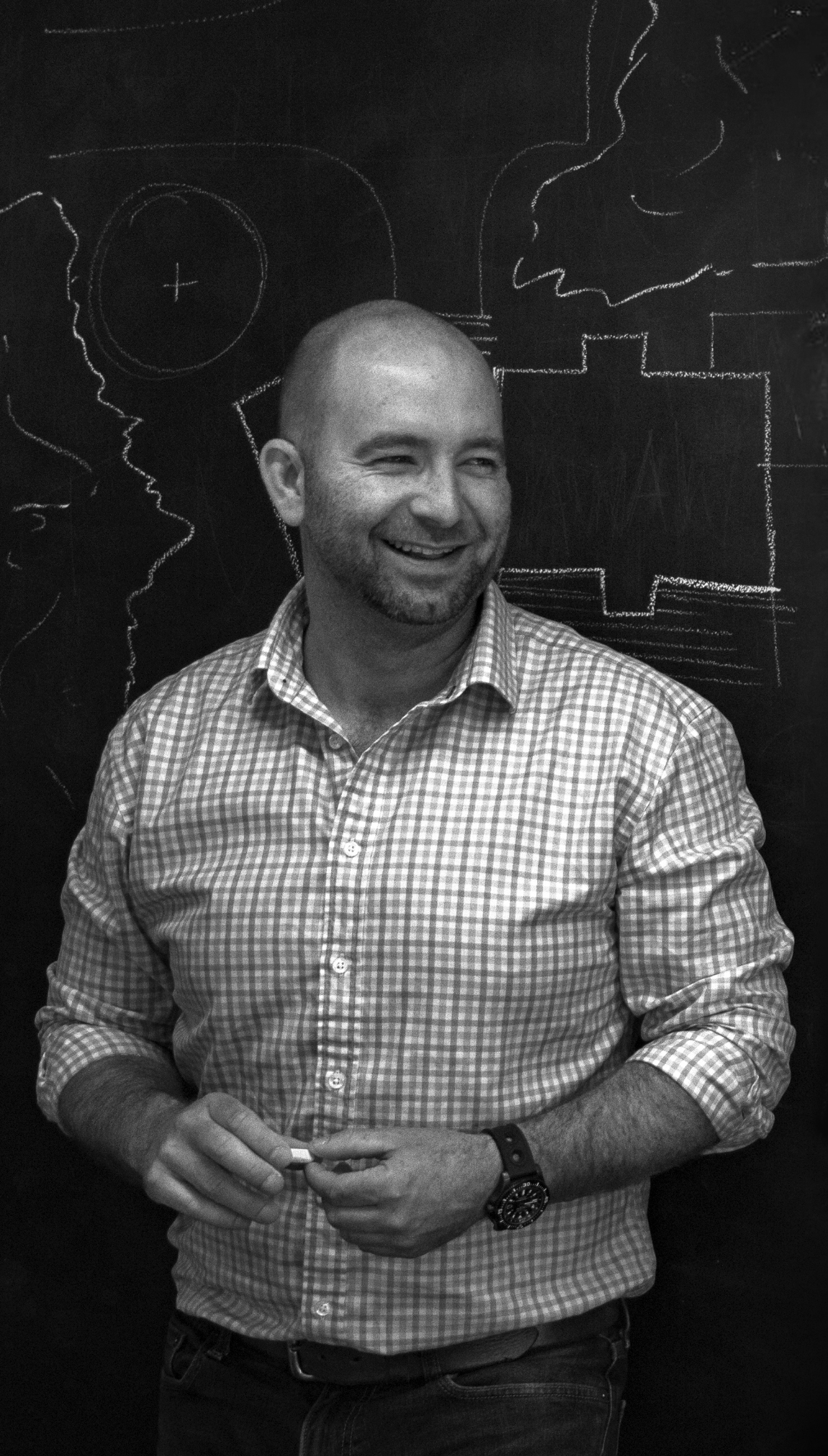 A man smiling and holding chalk in front of a blackboard with chalk drawings of faces and abstract shapes.