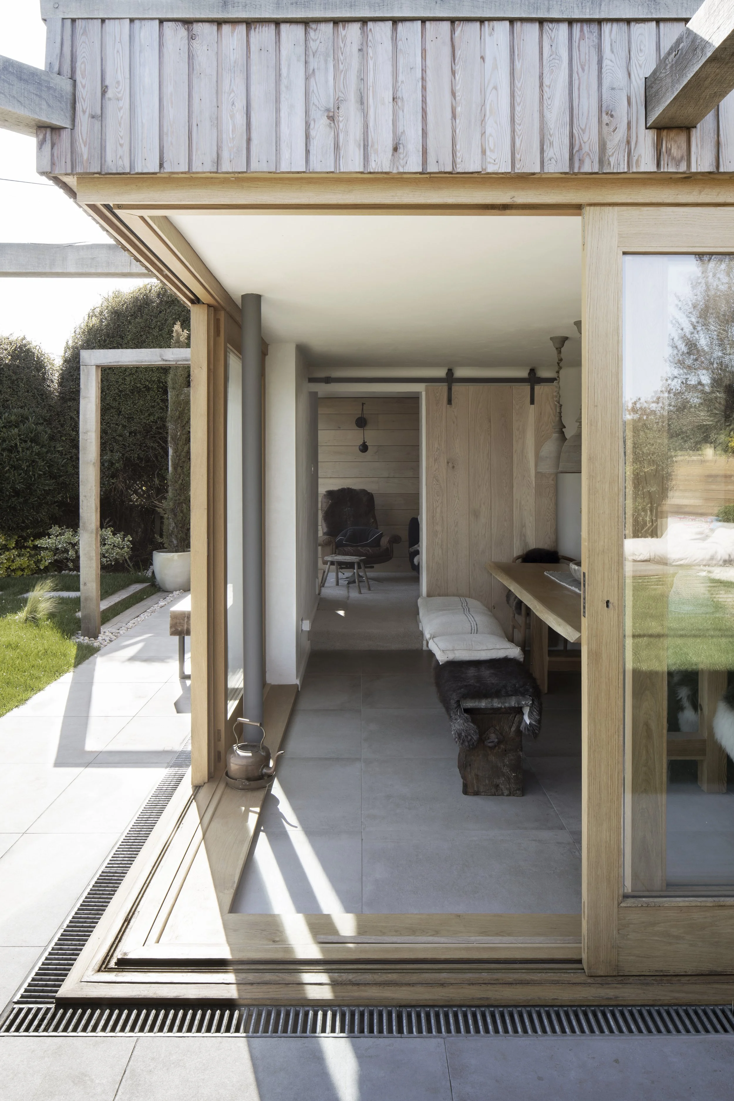 Interior view of a modern house with a wooden sliding door, a sitting area with chairs, a wooden bench with cushions, and a glimpse of a garden outside with grass and plants.