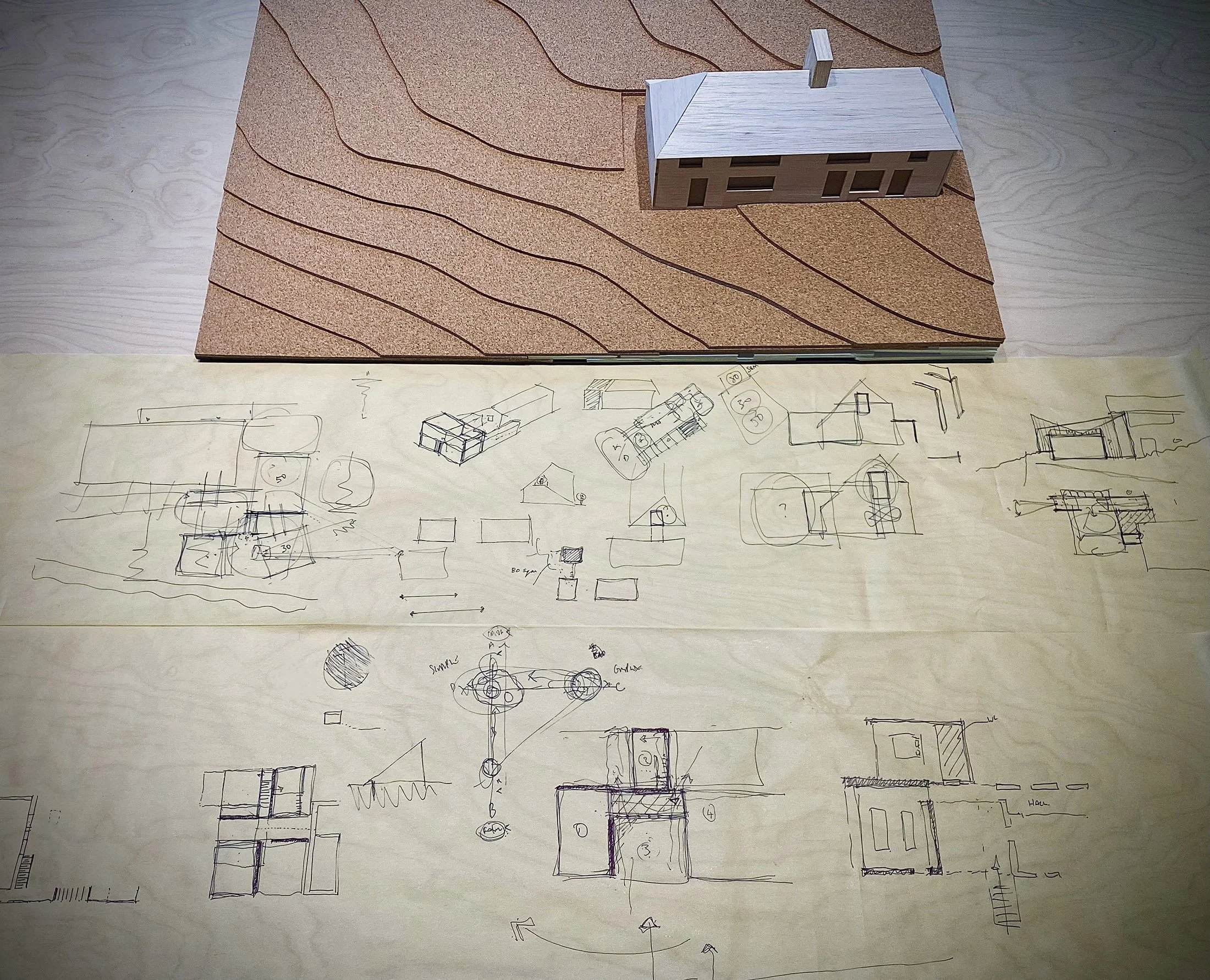 Paul Cashin Architects : A detailed architectural model of a house with a pitched roof, positioned on top of hand-drawn house plans and sketches on paper.