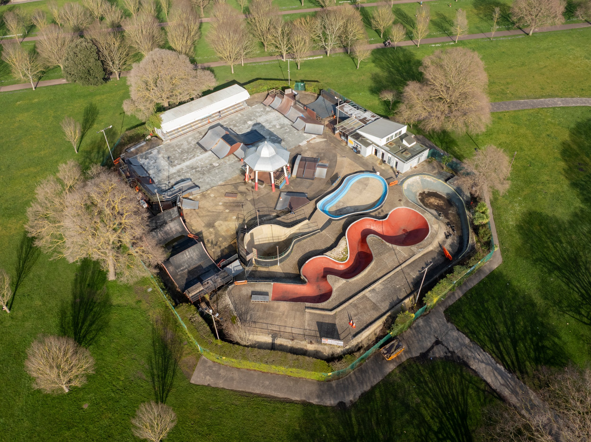 Aerial view of an empty outdoor skate park with multiple ramps, a small pool, and a water slide, surrounded by trees and grass.