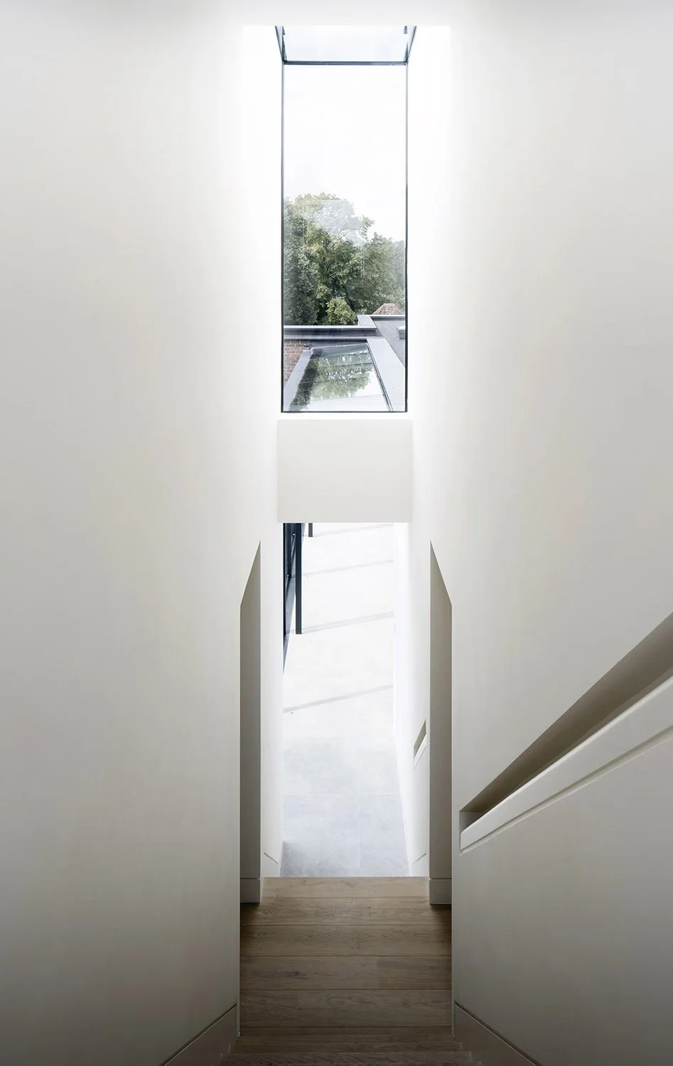 A view from a staircase looking up through a narrow, rectangular skylight window in a modern, white-walled interior.
