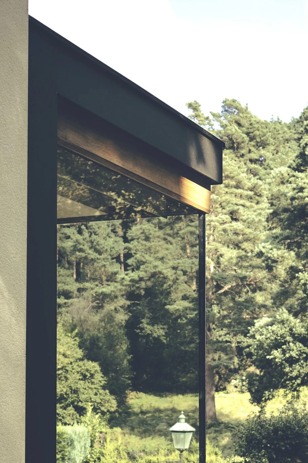 Close-up of a large window with black and wooden accents, overlooking a lush green forest with tall trees and a vintage lamp post in the background.