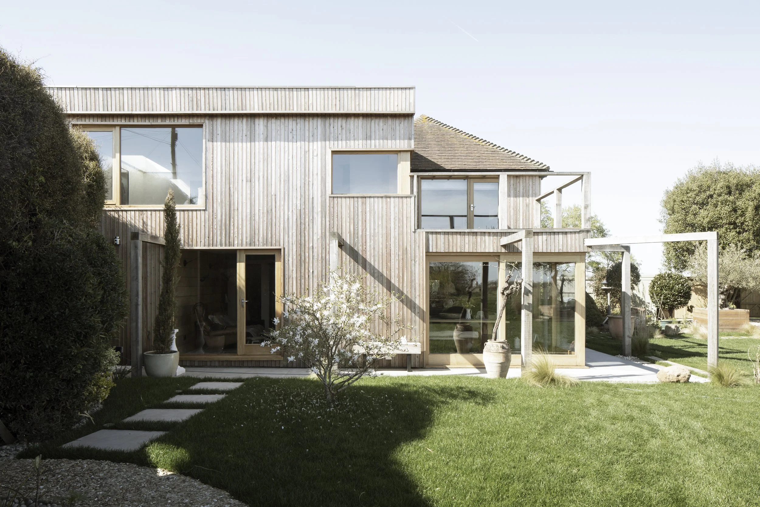 A modern two-story house with a wooden exterior, large windows, and a well-maintained garden with green grass, potted plants, and a small flowering tree.