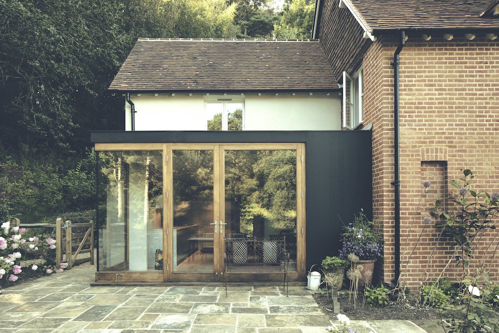 A modern house with a glass conservatory extension on a stone patio, surrounded by garden plants and flowers.
