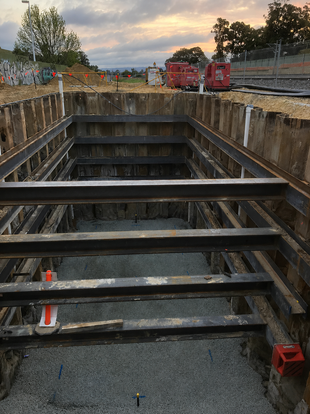 Construction site with excavated hole supported by steel beams, gravel base, and orange safety markers. In the background, there are construction equipment, fencing, and trees under a cloudy sky during sunset.
