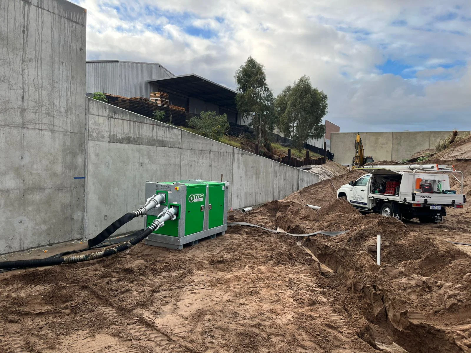 Construction site with a soil trench, construction equipment, a white utility truck, and a green machinery unit near a concrete wall, under a partly cloudy sky.