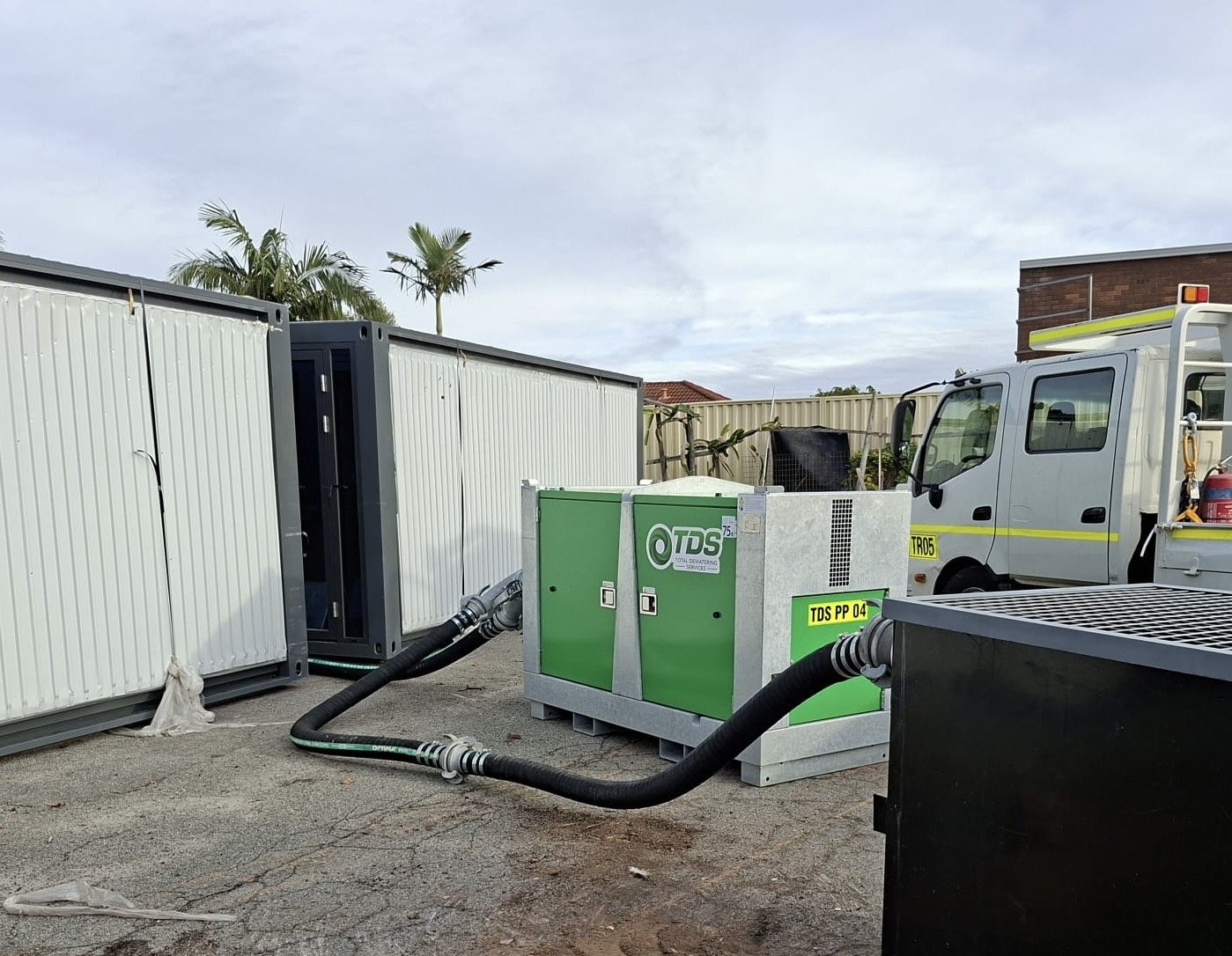 Outdoor scene with electrical equipment and machinery, including containers and a white utility vehicle with a yellow stripe, on a paved area with palm trees and a brick building in the background.