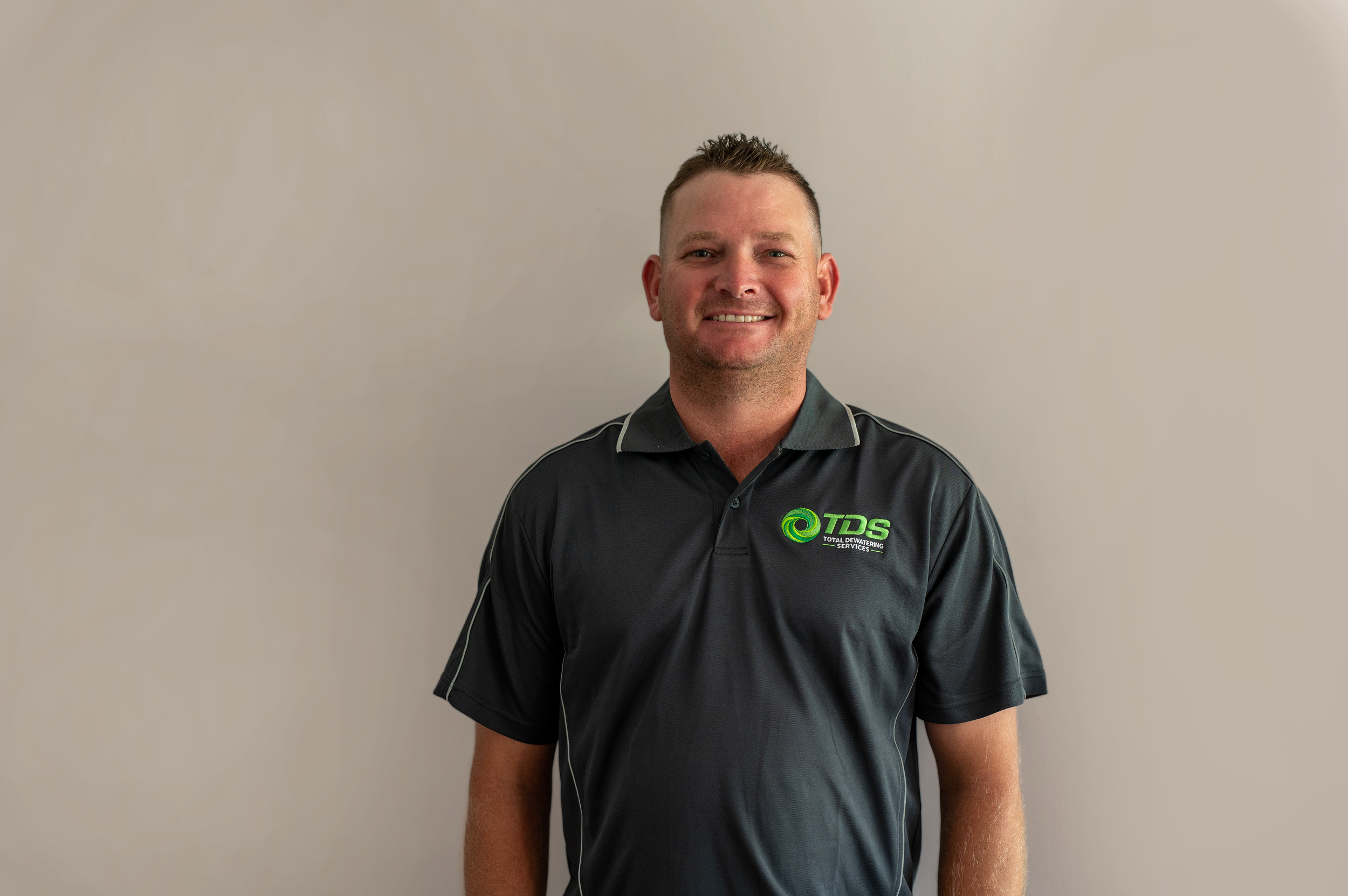Portrait of a smiling man in a black polo shirt with a logo, standing against a plain light-colored wall.