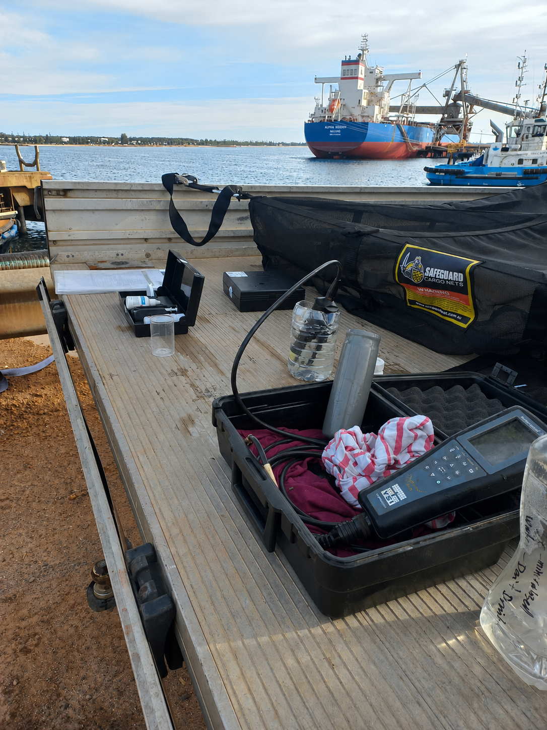 A workbench on a dock with scientific equipment, water bottles, and tools near the water, with boats and ships in the background.