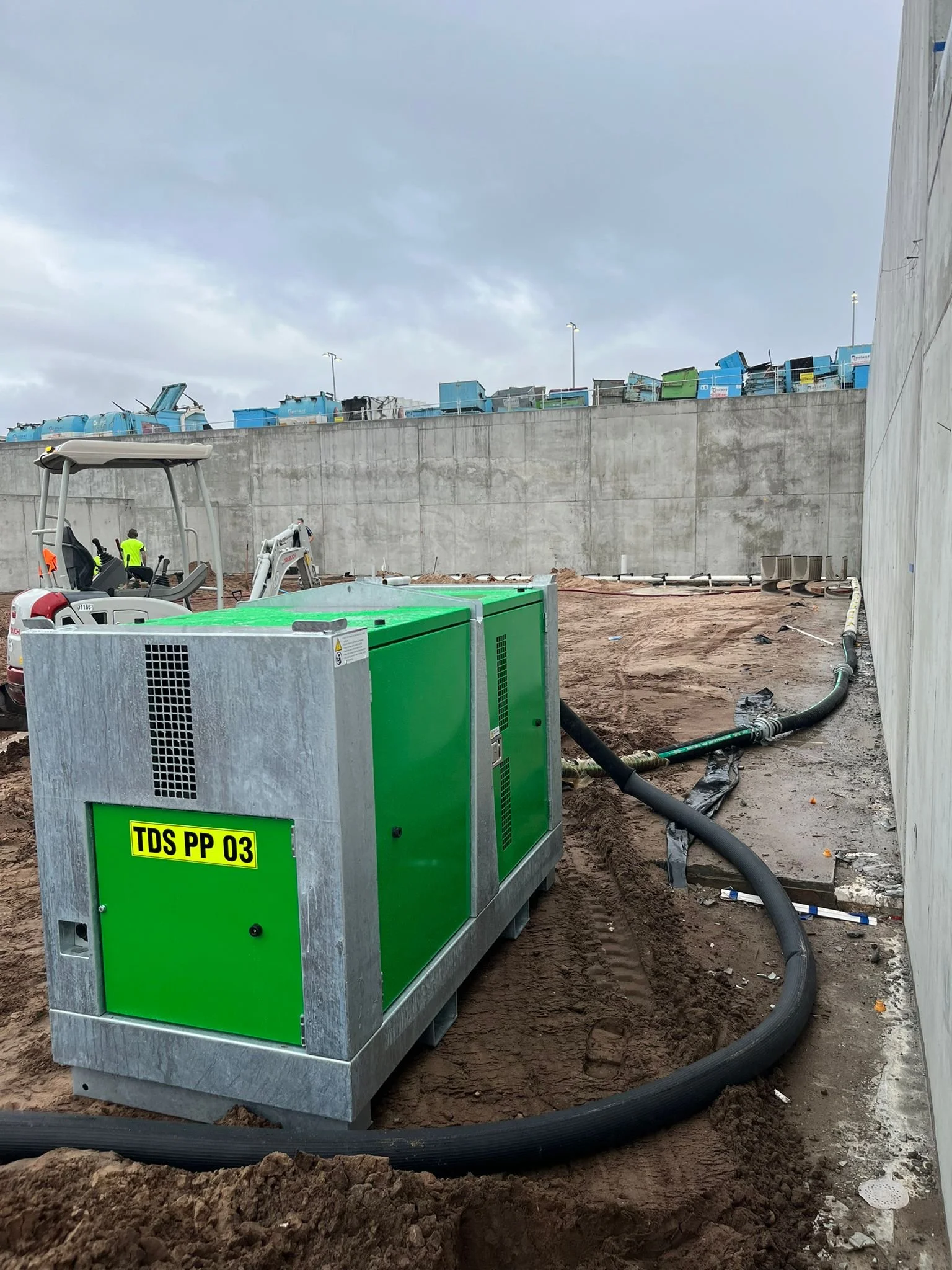 Construction site with large green HVAC units, dirt ground, black pipes, and workers in yellow reflective vests. Cloudy sky.