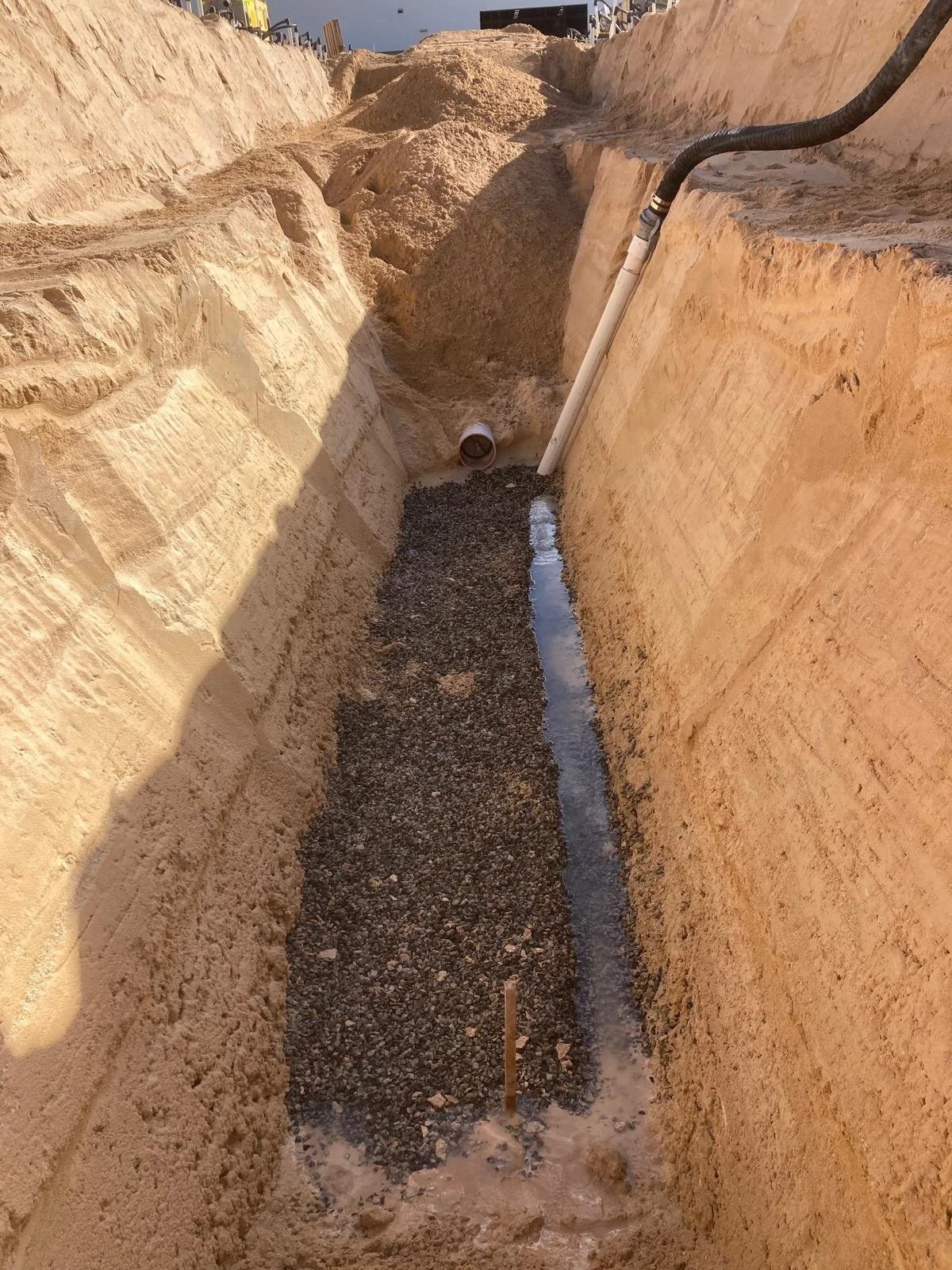 A construction trench with a drain pipe, gravel, and a pour concrete base, with excavated soil and construction equipment around.